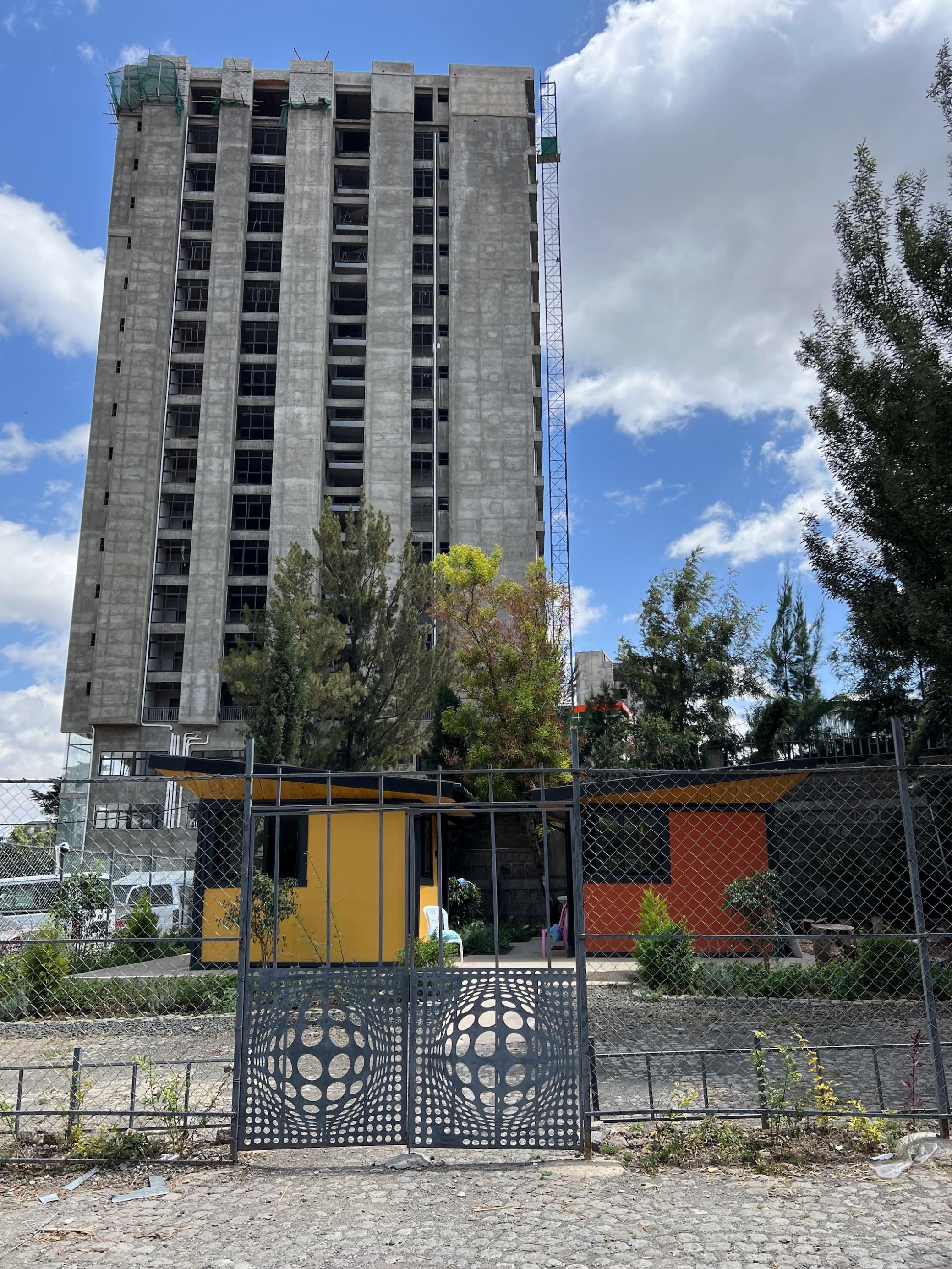 Vor einem Hochhaus im Rohbau stehen zwei kleine Häuser, das eine gelb, das andere orange gestrichen. Sie stehen hinter einem Zaum, auf dem Grundstück wächst etwas Grün.