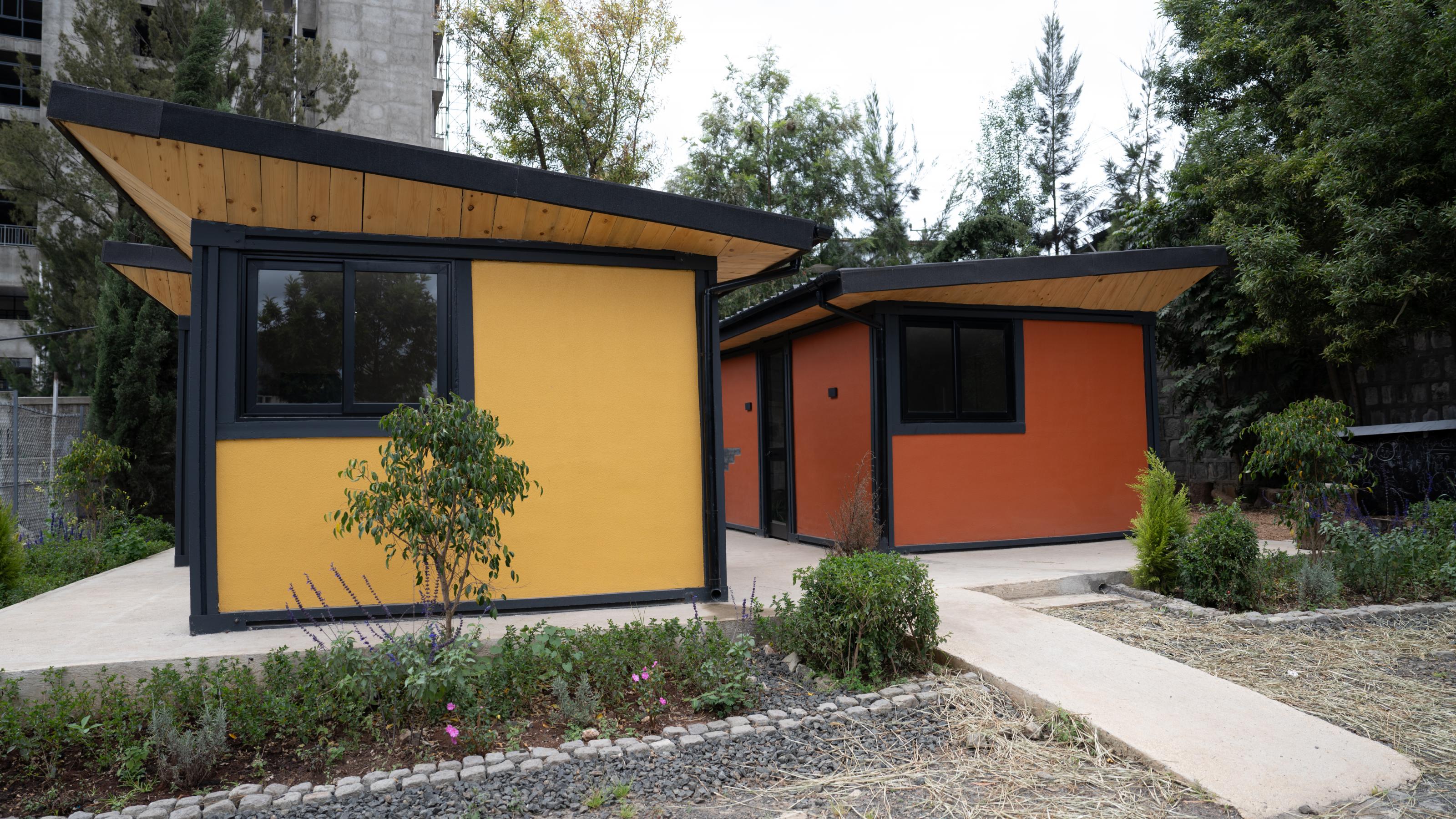 Zwei kleine Häuser mit Pultdächern, gelb und orange gestrichen. Im Hintergrund ist noch im Anschnitt ein Hochhaus zu sehen.