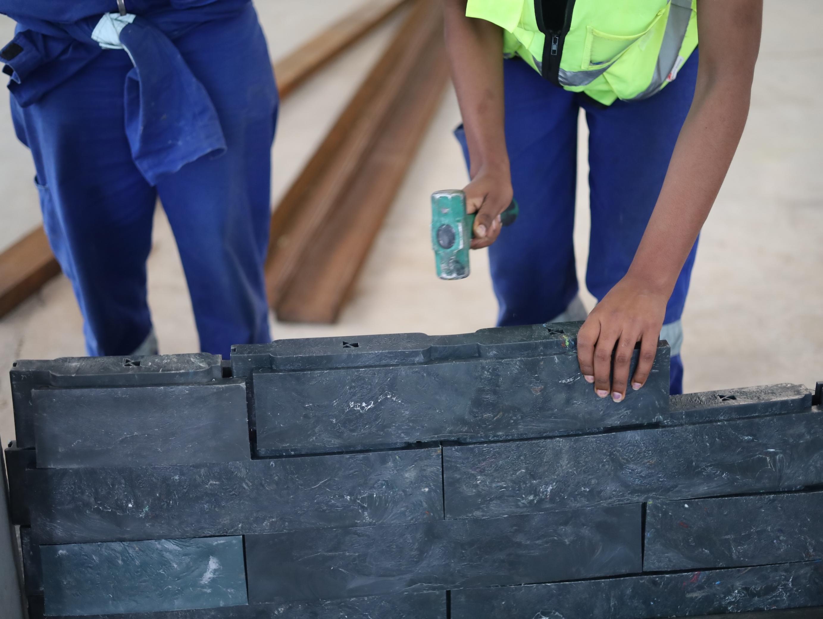 Nahaufnahme, die gräulichen Plastikziegel sind zu sehen, außerdem ein Hammer, der noch in der Luft schwebt und gleich auf sie niedergehen wird. Es ist zu erkennen, dass die Ziegel etwa wie Legosteine ineinander Halt finden.