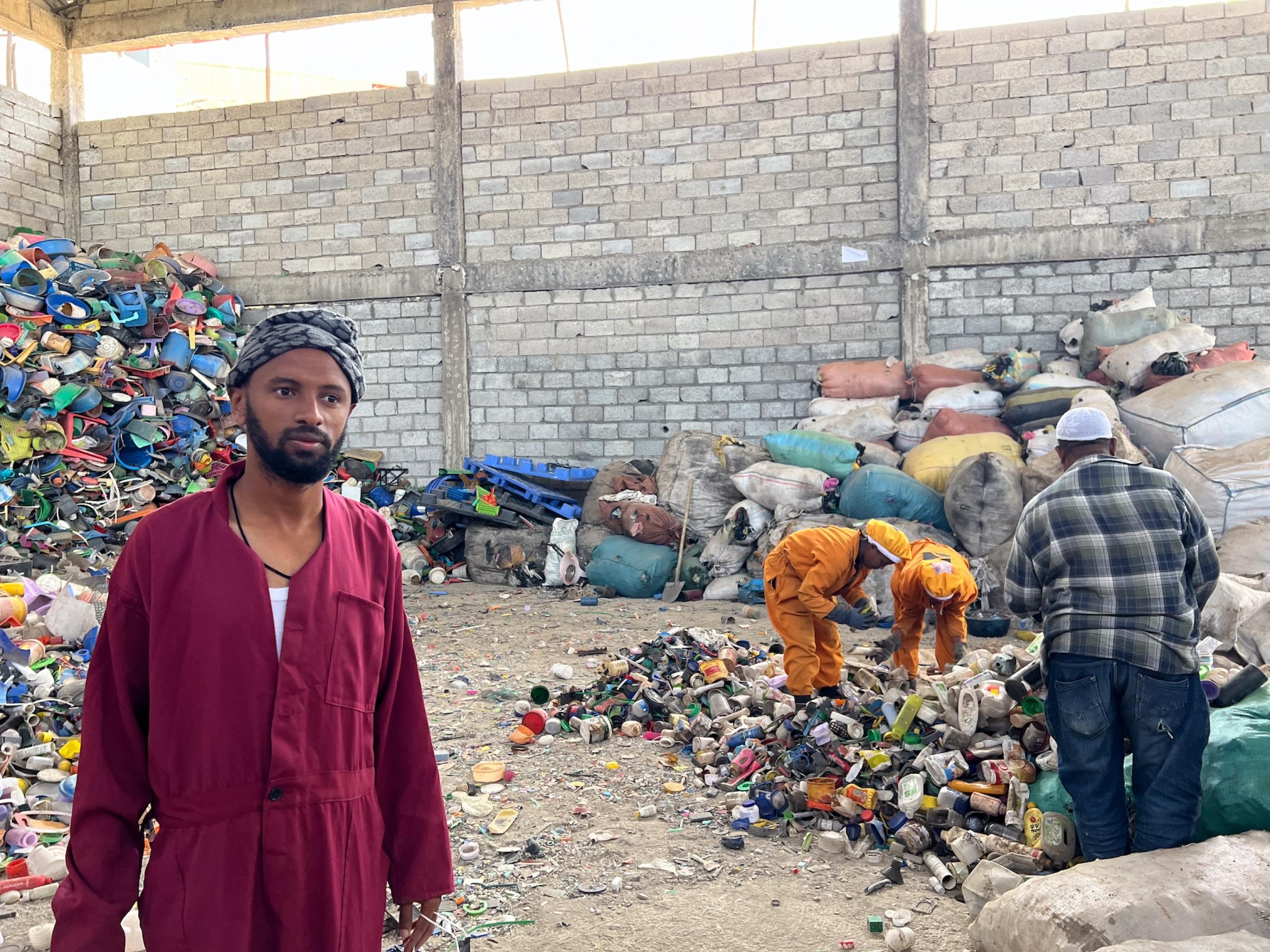 Eine Lagerhalle voller Plastikmüll, dazwischen ein Arbeiter und zwei Arbeiterinnen. Alle drei tragen Overalls als Arbeitskleidung.