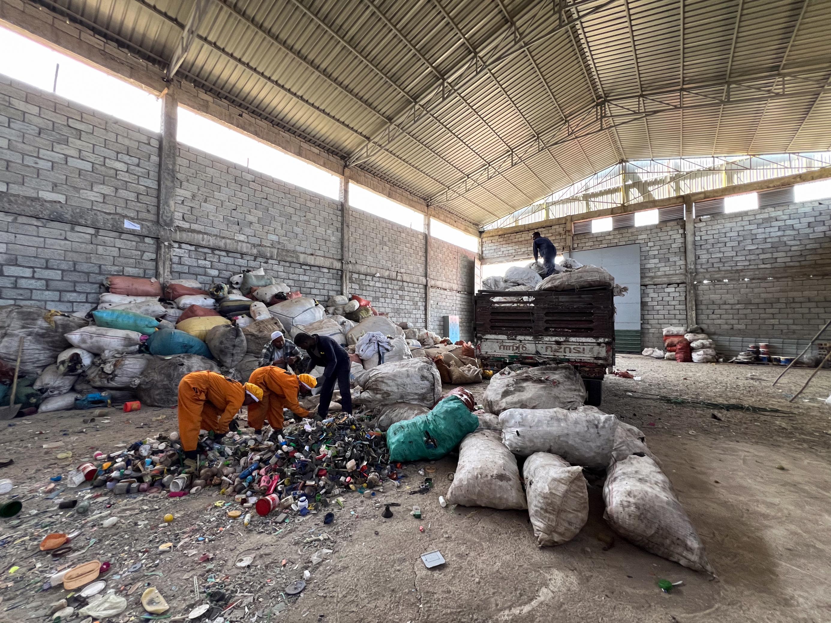 Eine Lagerhalle, gemauerte Wände. Im Vordergrund Müll und zwei Frauen sowie ein Mann in Arbeitskleidung, die ihn sortieren. Im Hintergrund ein LKW, die Ladefläche voller Säcke, einige sind schon abgeladen.