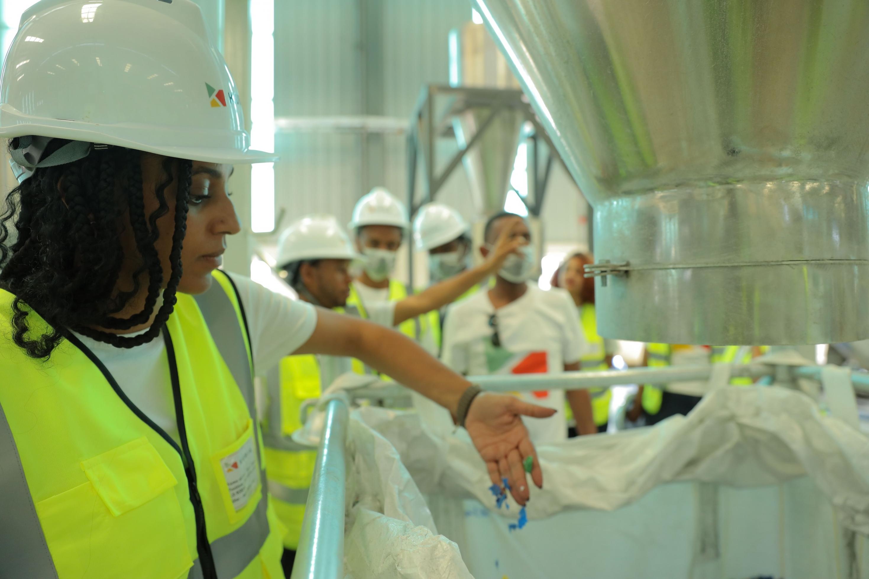 Eine Frau mit Helm und Signalweste steht vor einem Sack, aus dem sie offensichtlich die Flocken von buntem lastik geholt hat, die sie in der Hand hält.  hat sie einige