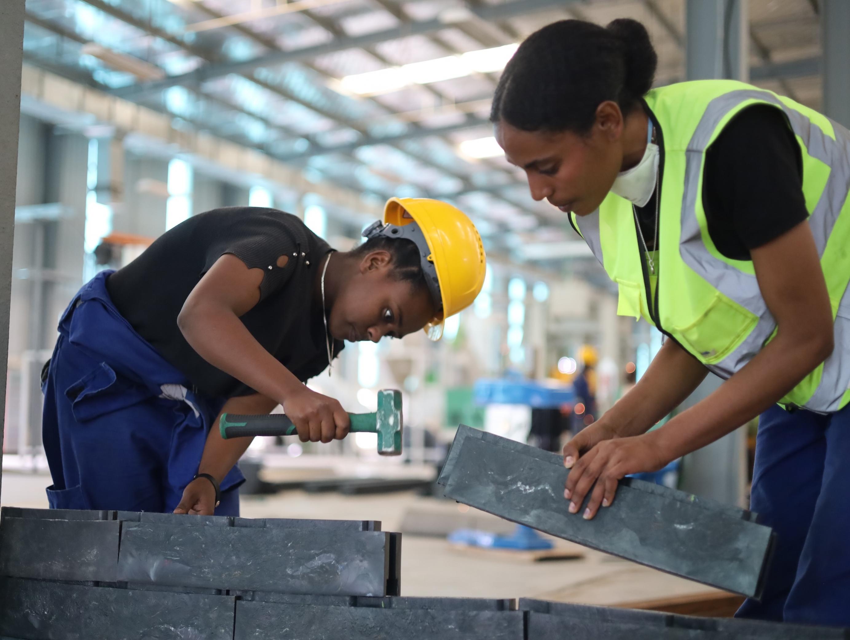 Zwei Frauen, beide tragen einen Helm, beugen sich über gräuliche Ziegel. Eine hat den Hammer erhoben, schlägt die Ziegel offenbar fest, die andere hält ihr schon den nächsten hin.