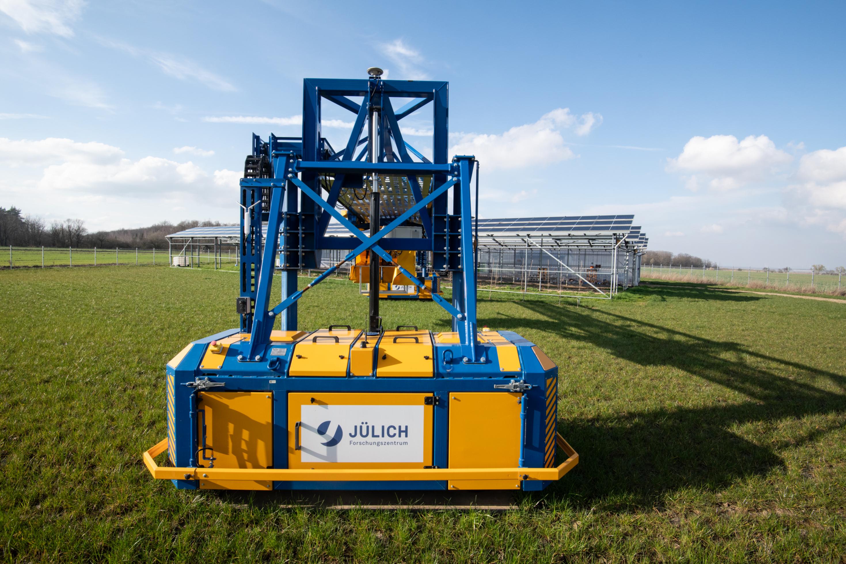 Im Vordergrund steht eine gelb-blaue Maschine auf einem Acker, im Hintergrund sind Metallgerüste mit Photovoltaikmodulen zu sehen.