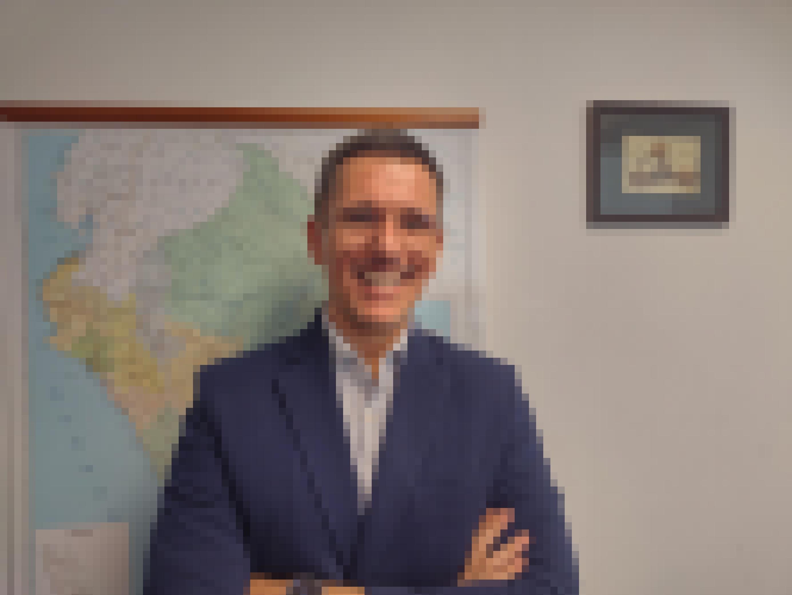 Ca. 40-jähriger Mann, schmales Gesicht, weiss, mit Brille, blaues Sakko, steht vor einer Landkarte in einem Büro und blickt in die Kamera