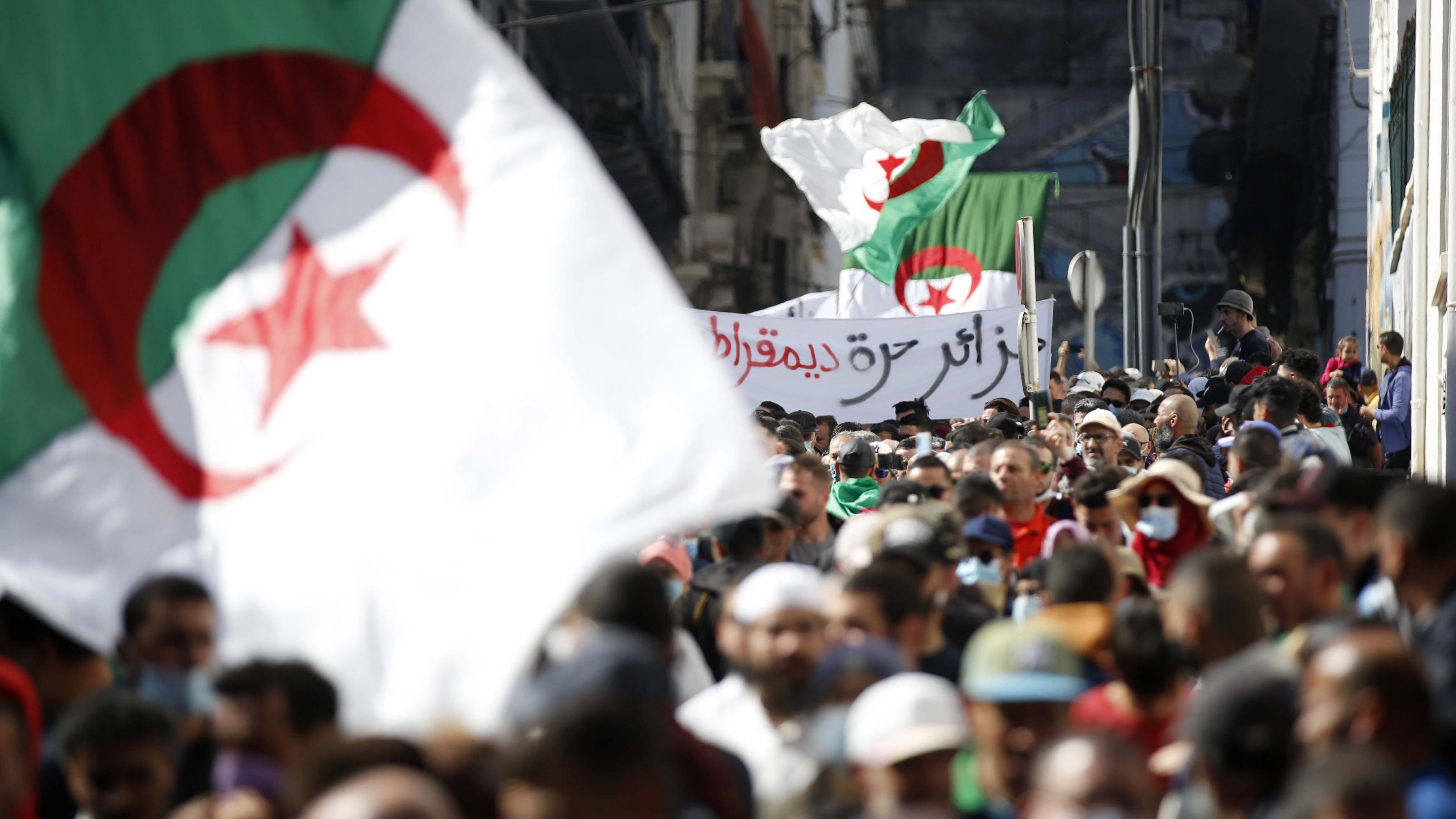 Demonstranten in einer engen Straße in Algier schwenken große algerische Fahnen. Im Hintergrund ist ein arabischsprachiges Transparent zu sehen.