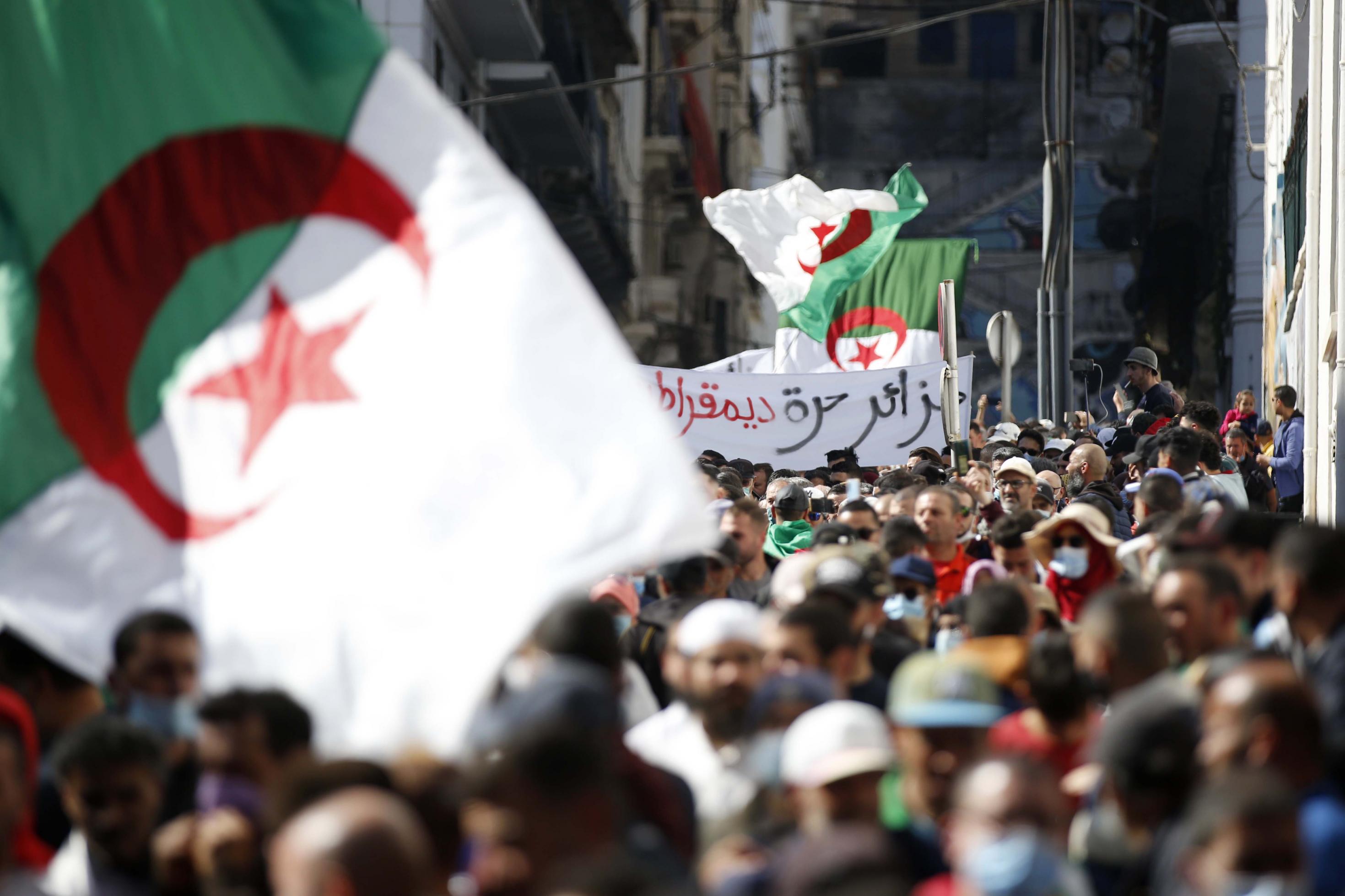 Demonstranten in einer engen Straße in Algier schwenken große algerische Fahnen. Im Hintergrund ist ein arabischsprachiges Transparent zu sehen.