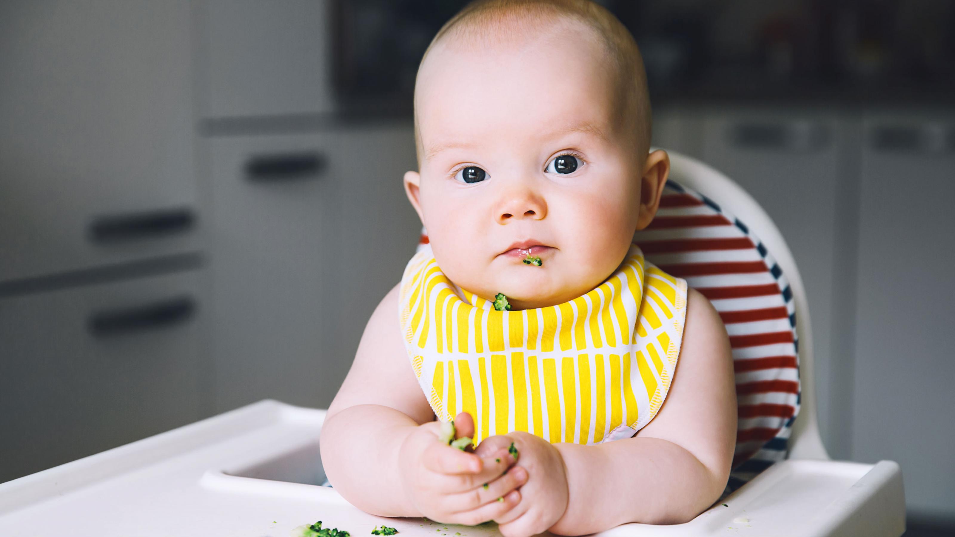 Ein kleines Kind sitzt in seinem Hochstuhl mit einem Lätzchen um und isst Brokkoli
