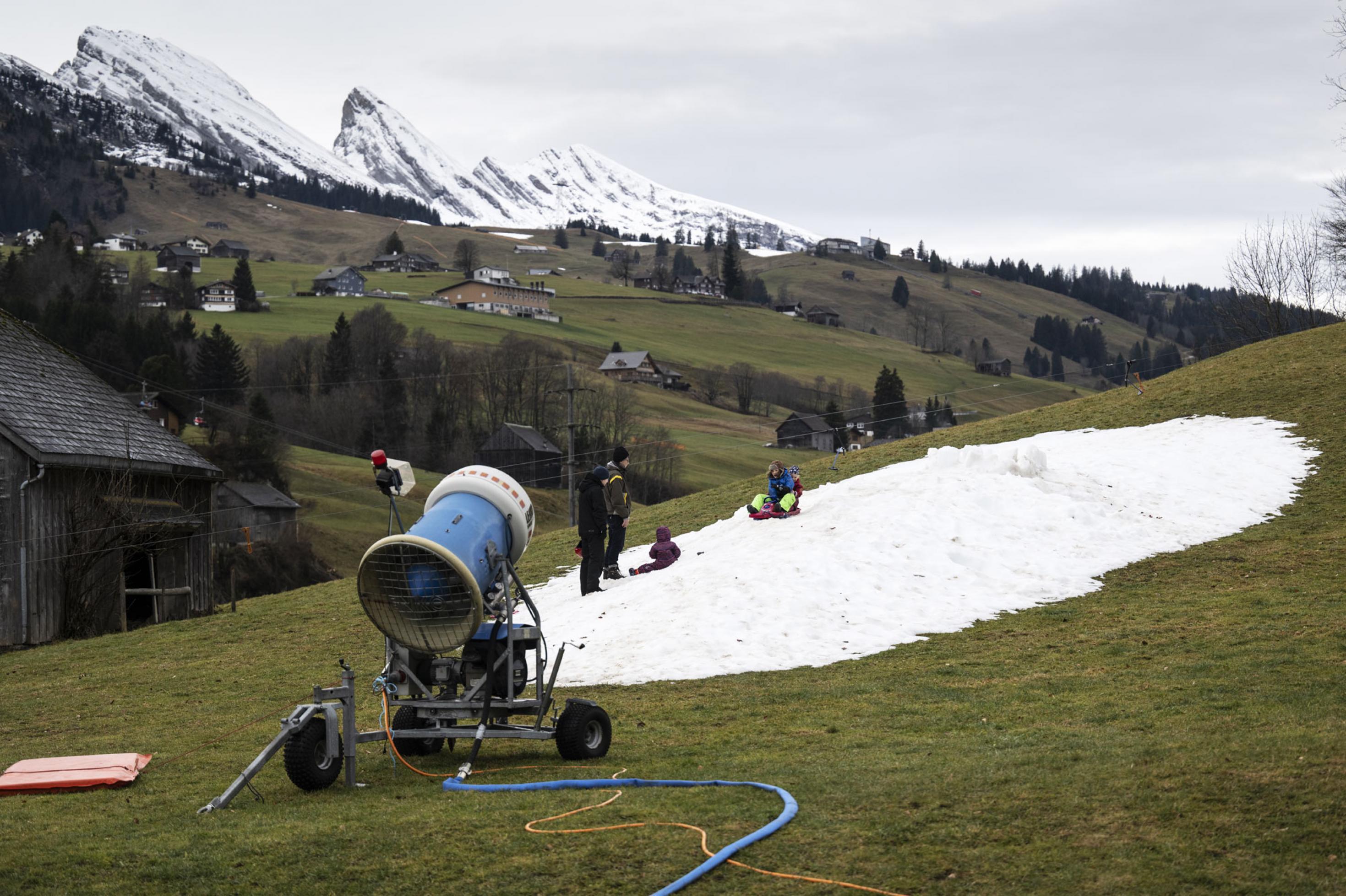 Eine Schneekanone vor einem kleinen Fleck Schnee, auf dem Kinder spielen, darum grüne Wiesen vor Bergkulisse.