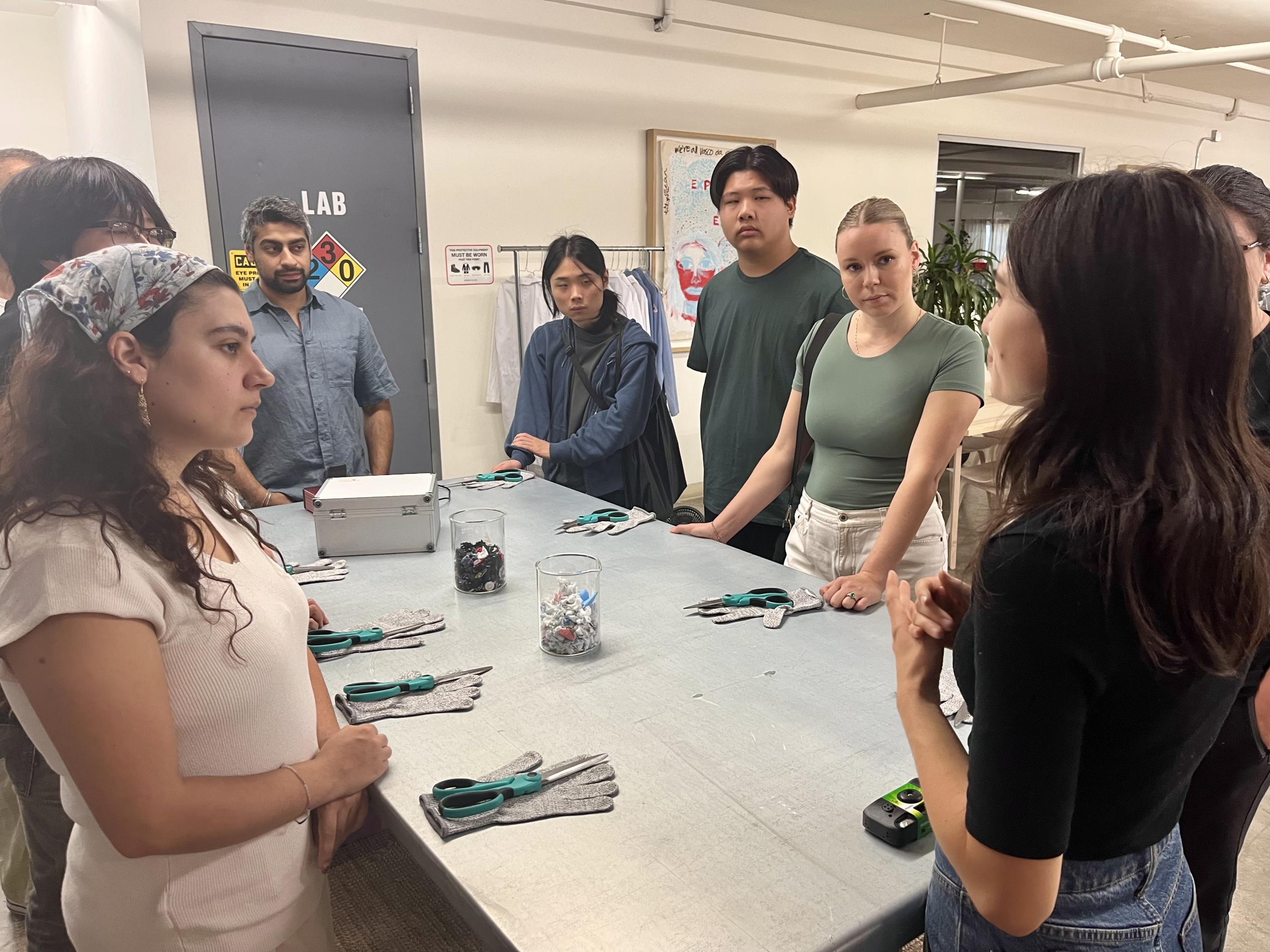 Studentinnen und Studenten stehen um einen Tisch mit Scheren und Handschuhen, während eine Mitarbeiterin die Geschichte von Ambercycle erklärt.