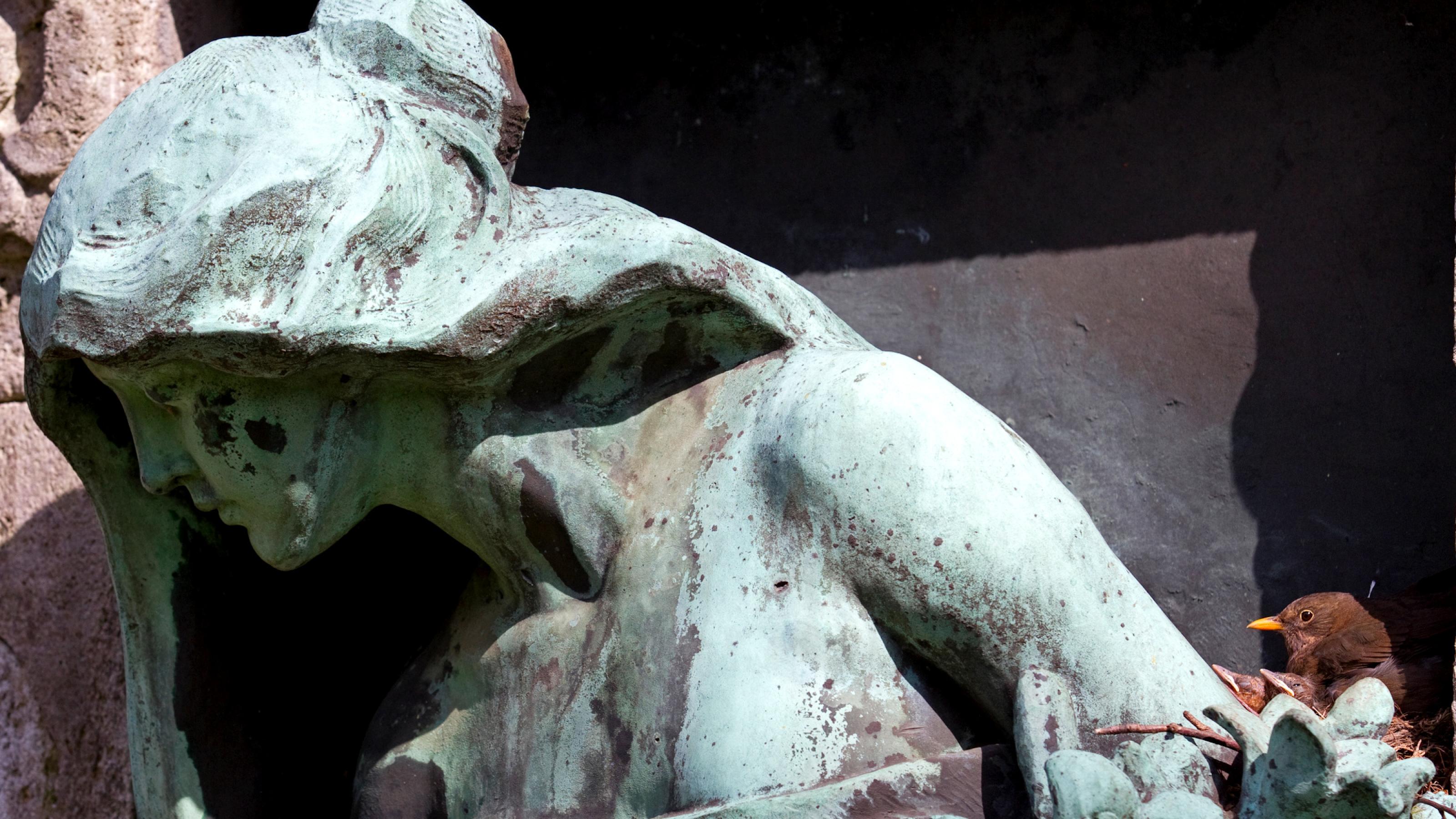 Zu sehen ist eine Bronzestatue in Frauengestalt. Ganz rechts unten in der der Ecke, auf einem Vorsprung der Statue, ist ein Nest, auf dem ein brauner Vogel sitzt.