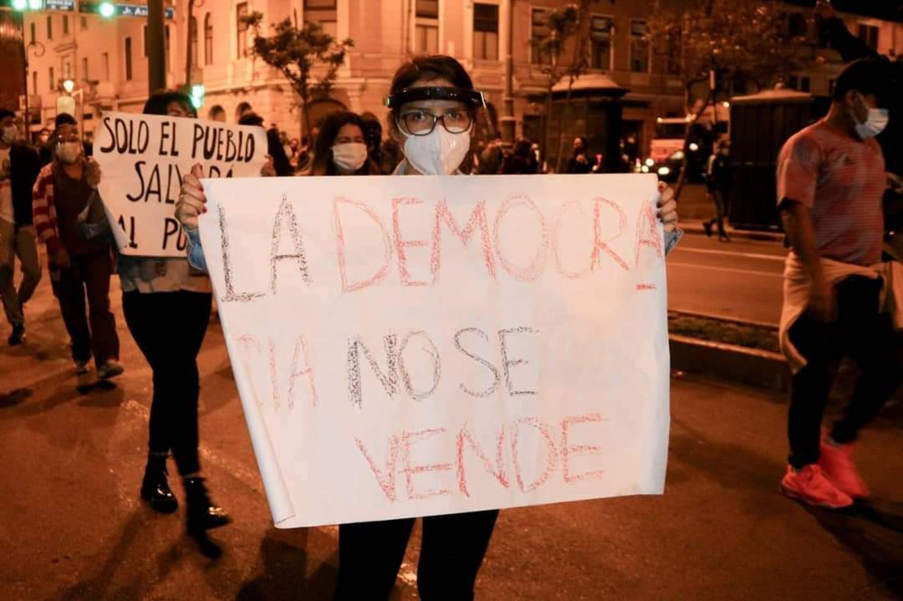 Junge Frau mit weißem Mundschutz, Brille, trägt ein Plakat vor der Brust, auf dem in spanisch steht: „La democracia no se vende“. Die Demokratie lässt sich nicht verkaufen.