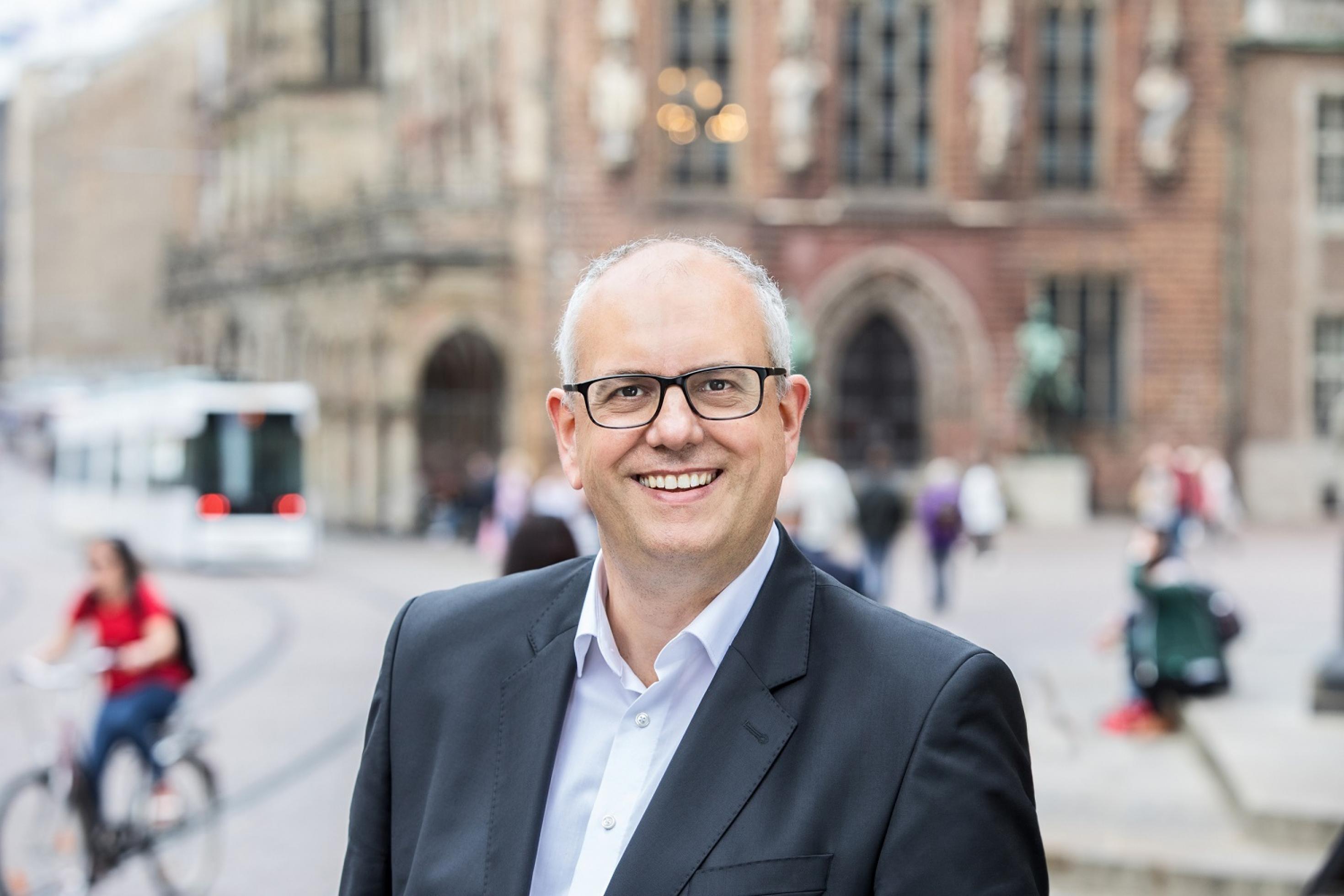 Ein Mann mit Brille und Halbglatze lächelt in die Kamera, im Hintergrund das historische Rathaus von Bremen.