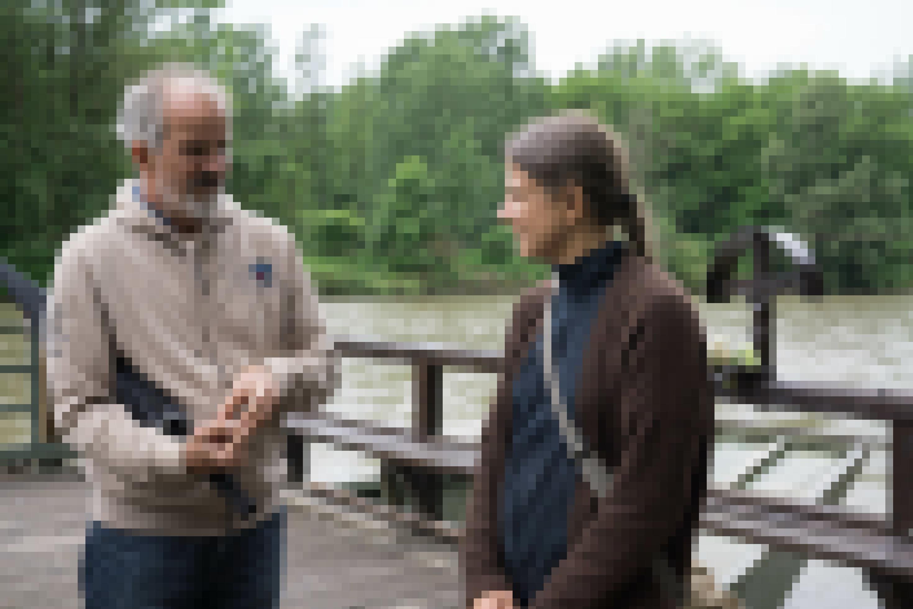 Mann mit grauem Bart und Frau mit langem Zopf stehen auf Holzplattform am Wasser und unterhalten sich.