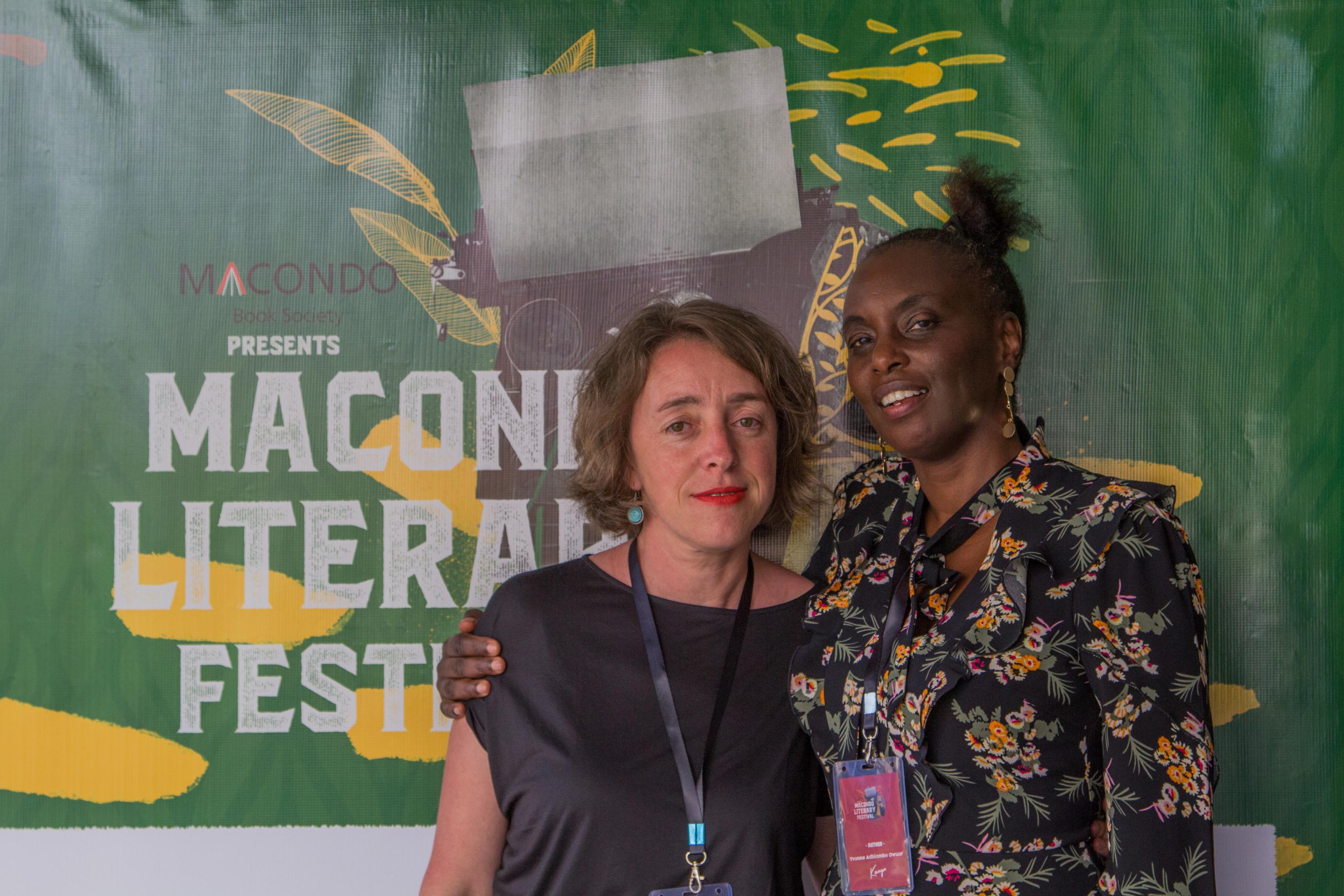 Zwei jüngere Frauen stehen vor einer Plakatwand. Auf dem Plakat steht „Macono Literary Festival“.