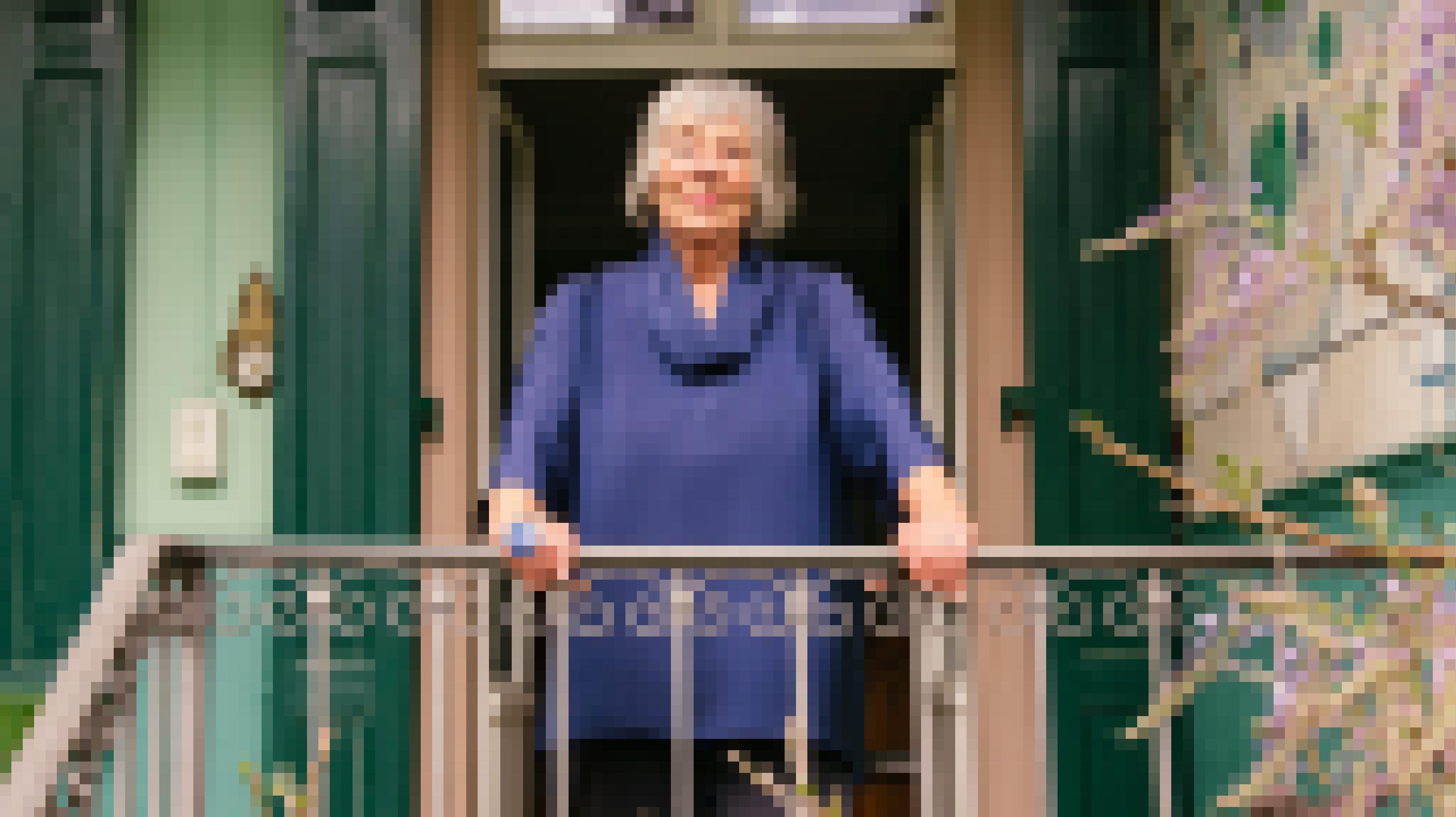 Eine sympathische ältere Dame mit grauen Haaren steht auf einem Balkon und lächelt in die Kamera.