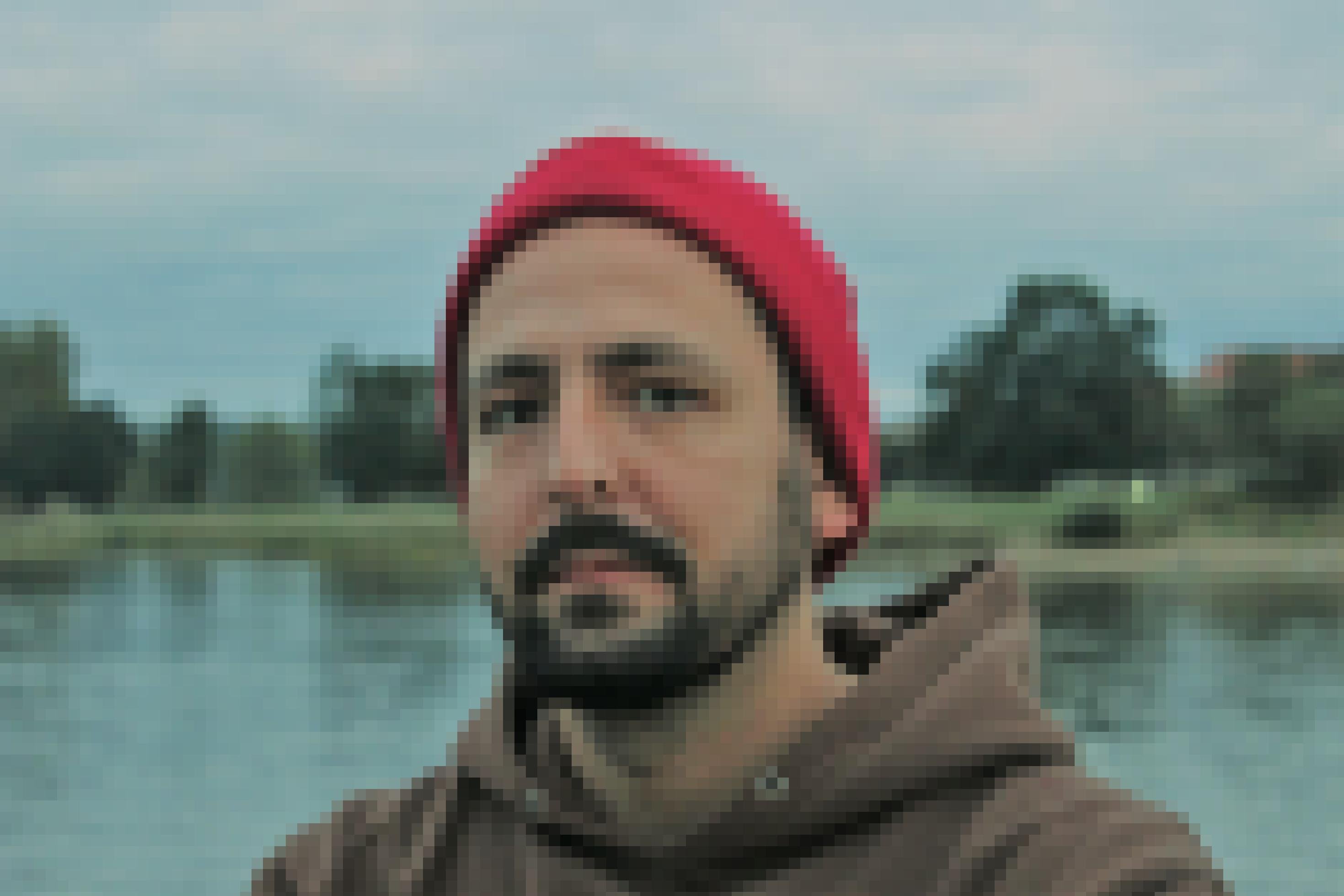 Jan Gehler guckt mit Bart, braunem Hoodie und roter Mütze in die Kamera. Im Hintergrund ein See, am anderen Ufer ein paar Bäume.