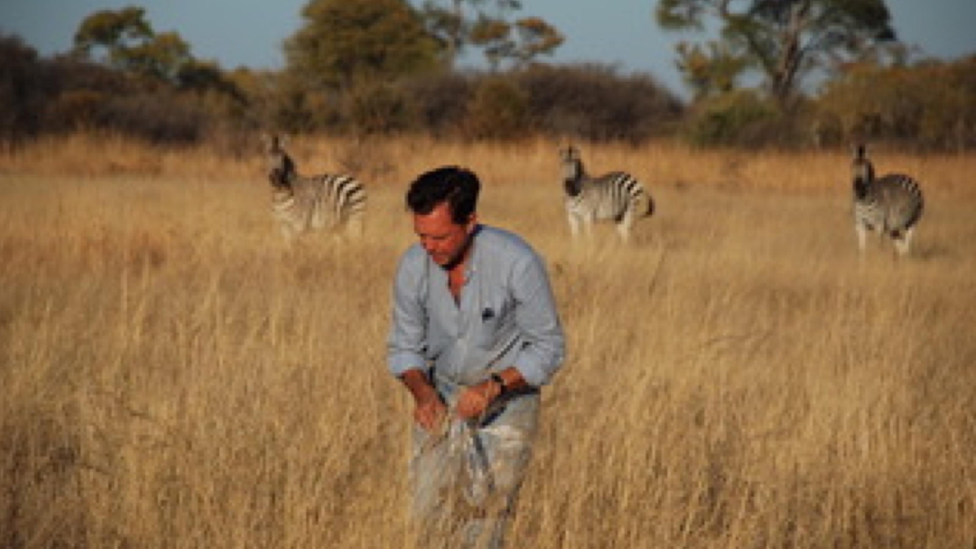 Patrick Duncan steht in hohem, trockenem Gras. Im Hintergrund sind drei Zebras.