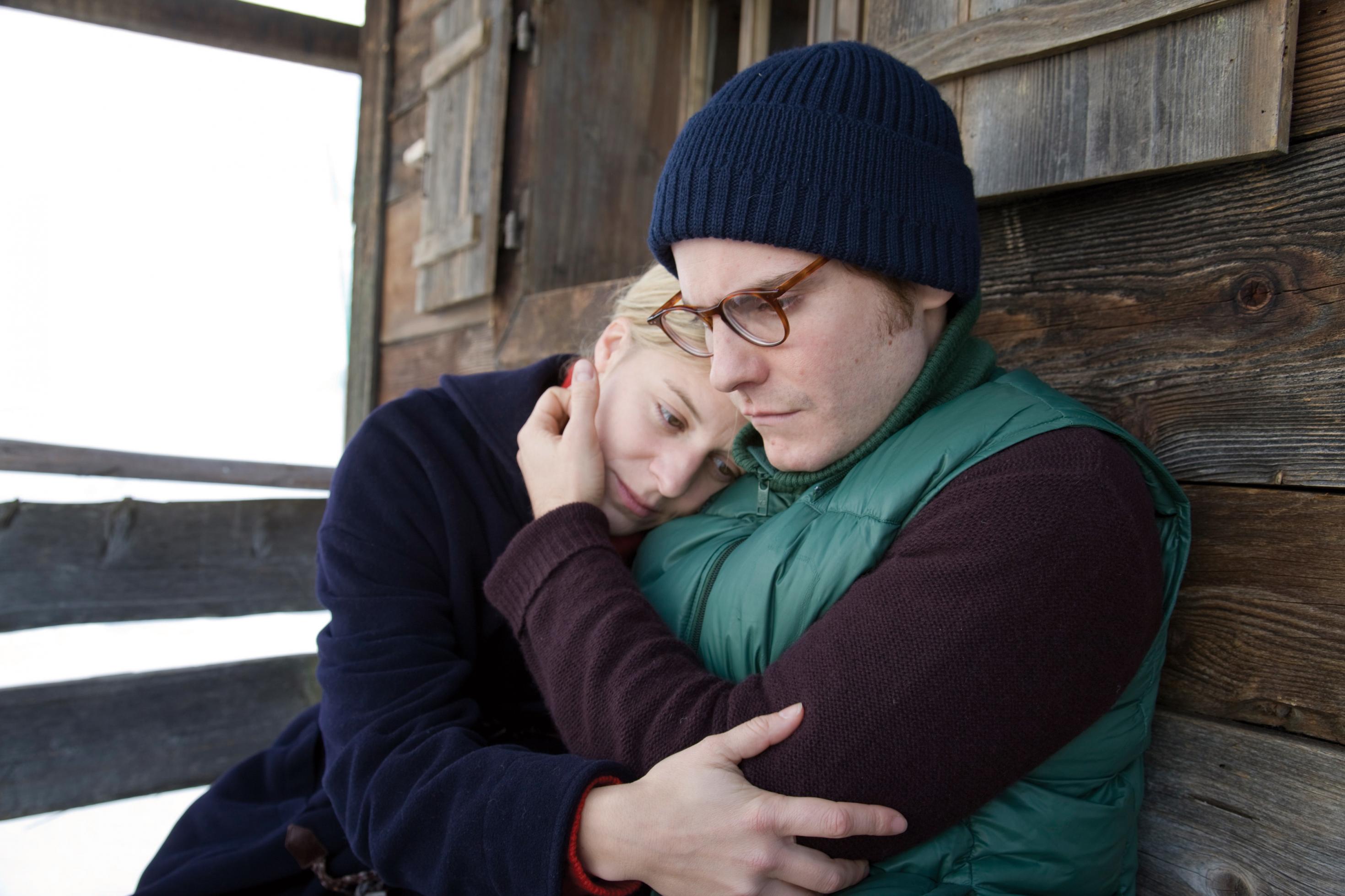 Laura Kuper (Bernadette Heerwagen) und Hans Krämer (Daniel Brühl) sitzen vor einer Holzhütte und halten sich gegenseitig im Arm.