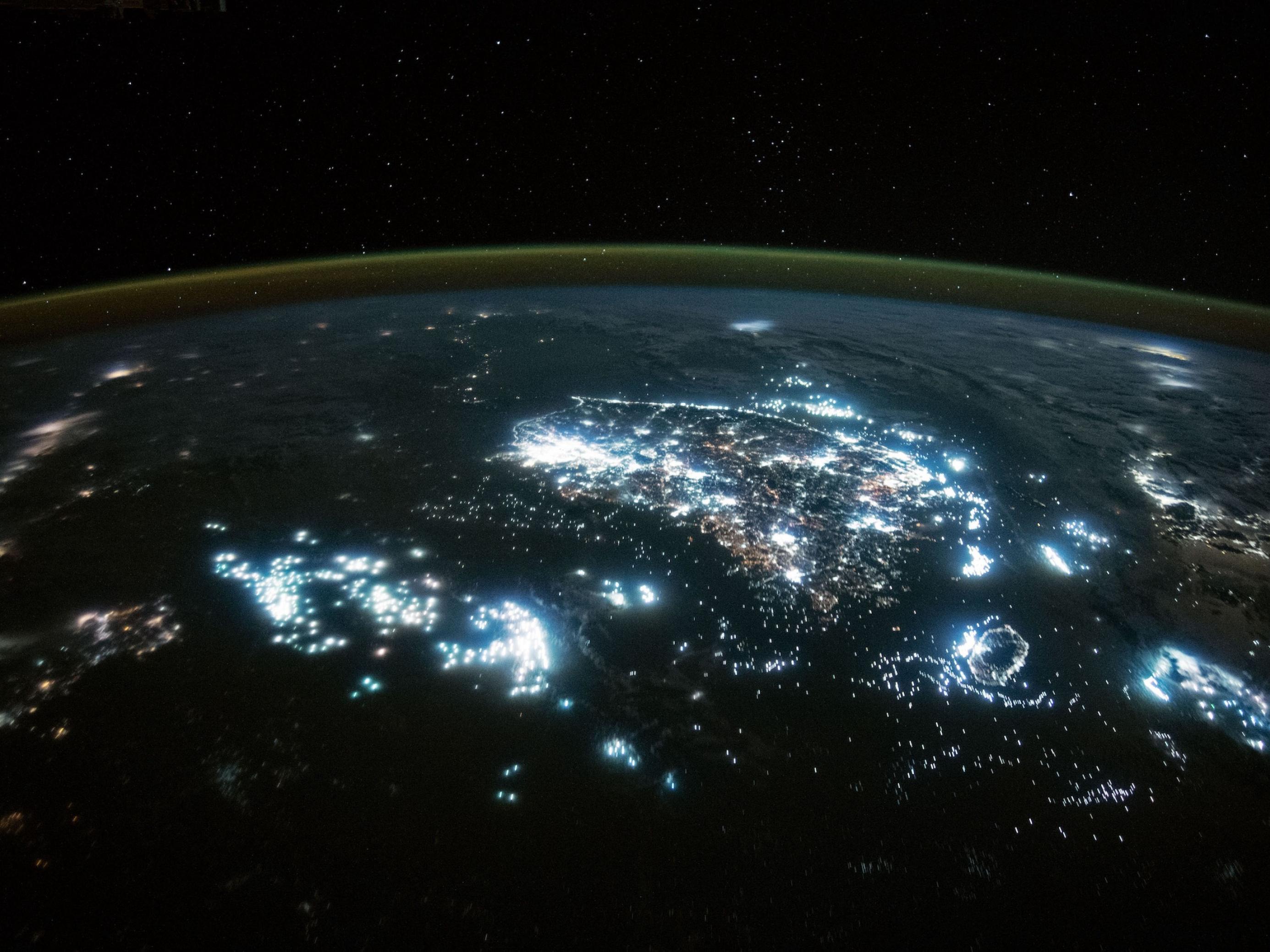 Nachtaufnahme vom Weltall aus, man sieht die gekrümmte Erdkugel und nächtliche Städte, die hell leuchten, sowie helle Flecken auf dem Meer, die Fischereiflotten.