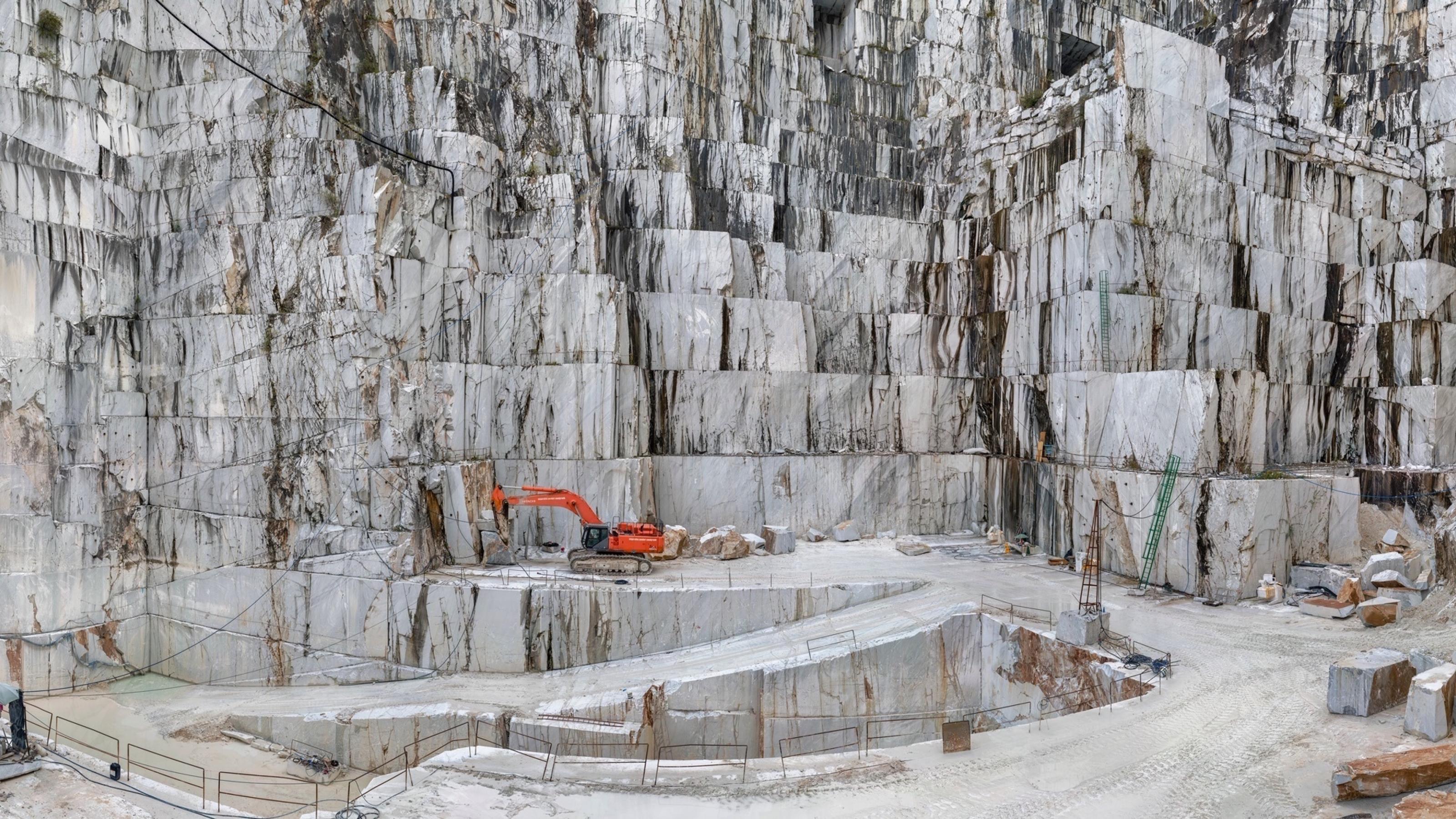 Dargestellt ist der Marmorsteinbruch von Carrara – vor der Kulisse riesiger weißer Felswände steht ein Bagger, mit dessen Hilfe Menschen die Gebirgslandschaft verändern.