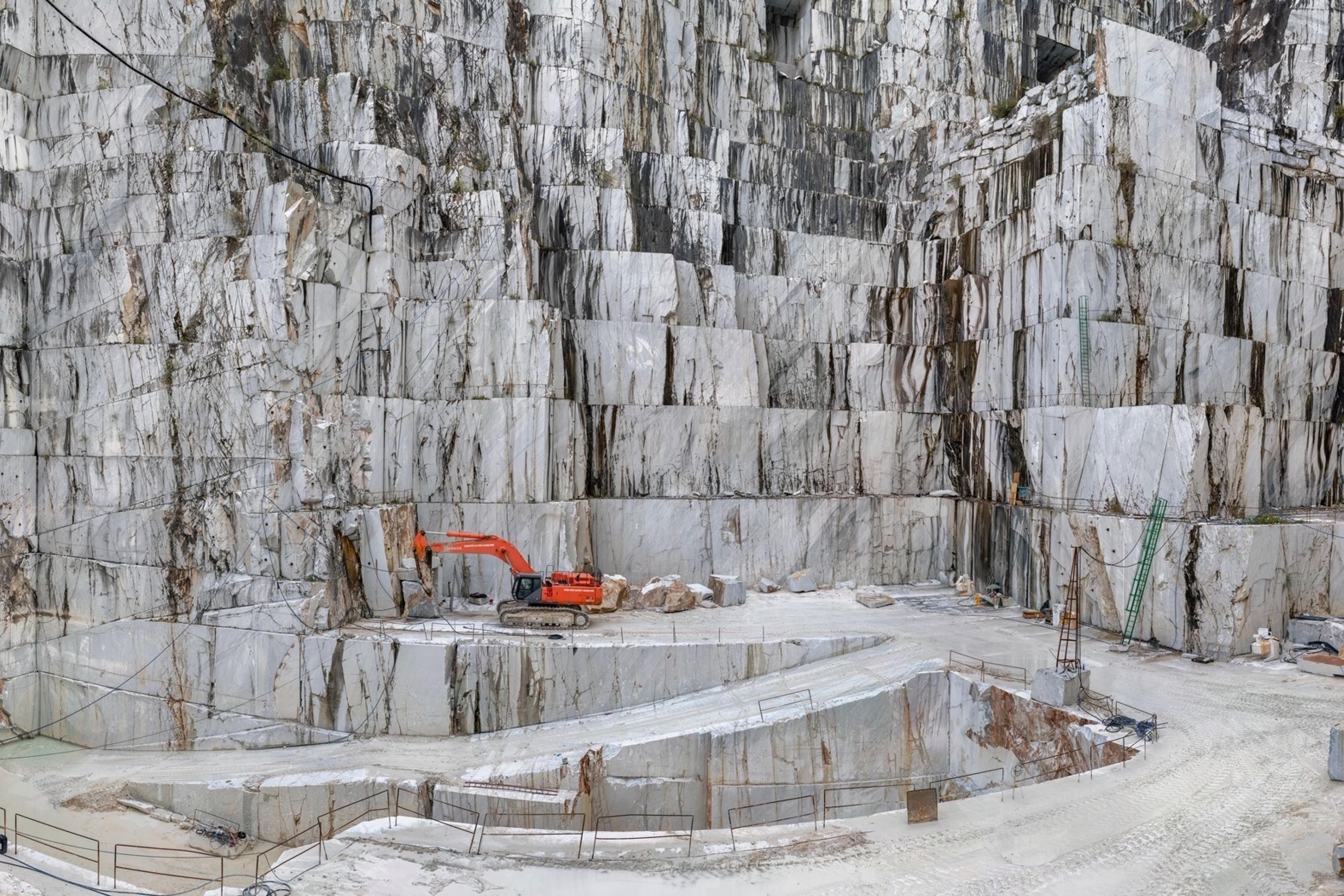 Dargestellt ist der Marmorsteinbruch von Carrara – vor der Kulisse riesiger weißer Felswände steht ein Bagger, mit dessen Hilfe Menschen die Gebirgslandschaft verändern.