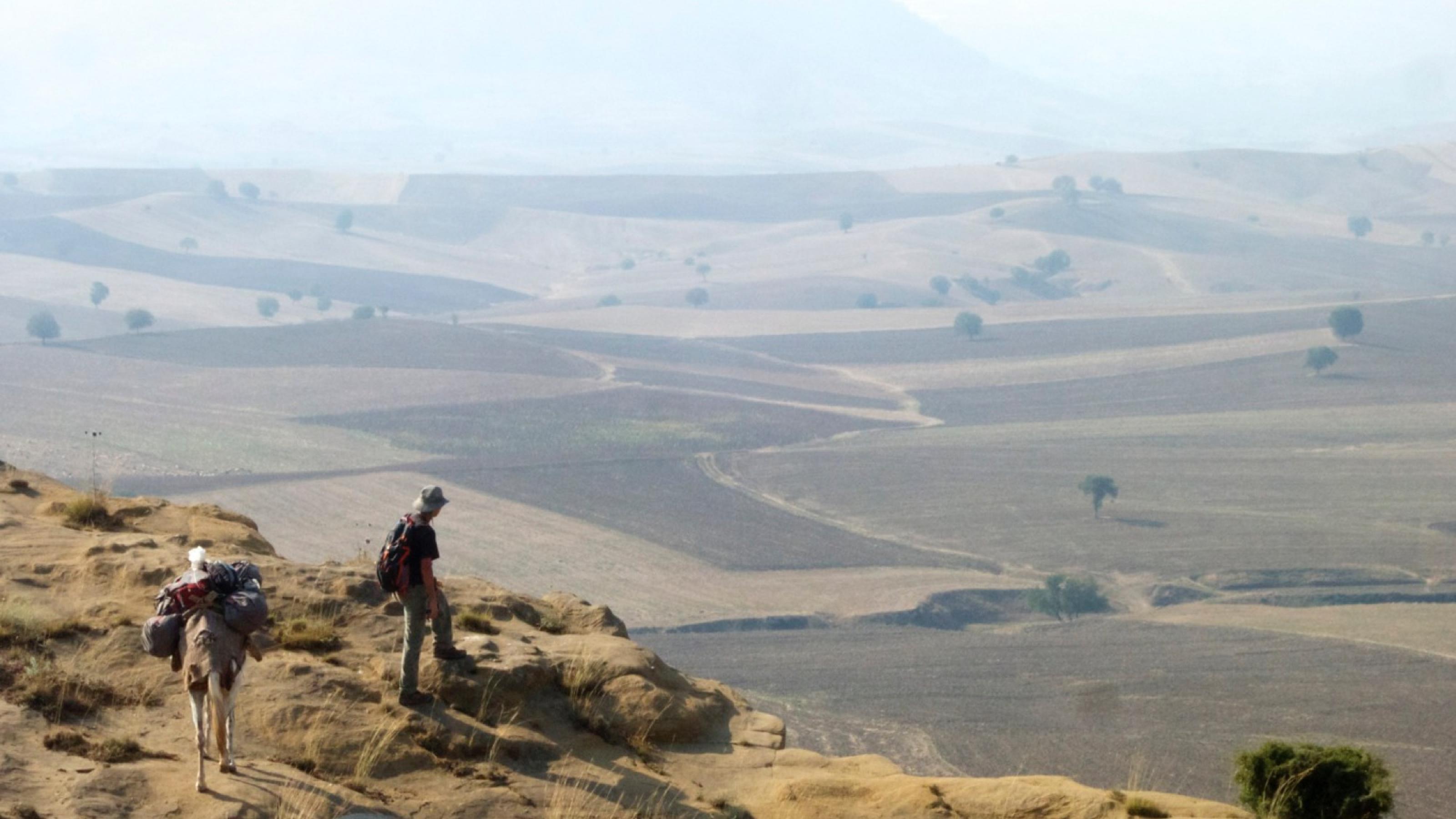 Der Journalist Paul Salopek steht am Rand eines Berges und schaut in die Ferne. Dort sind viele Felder zu erkennen.