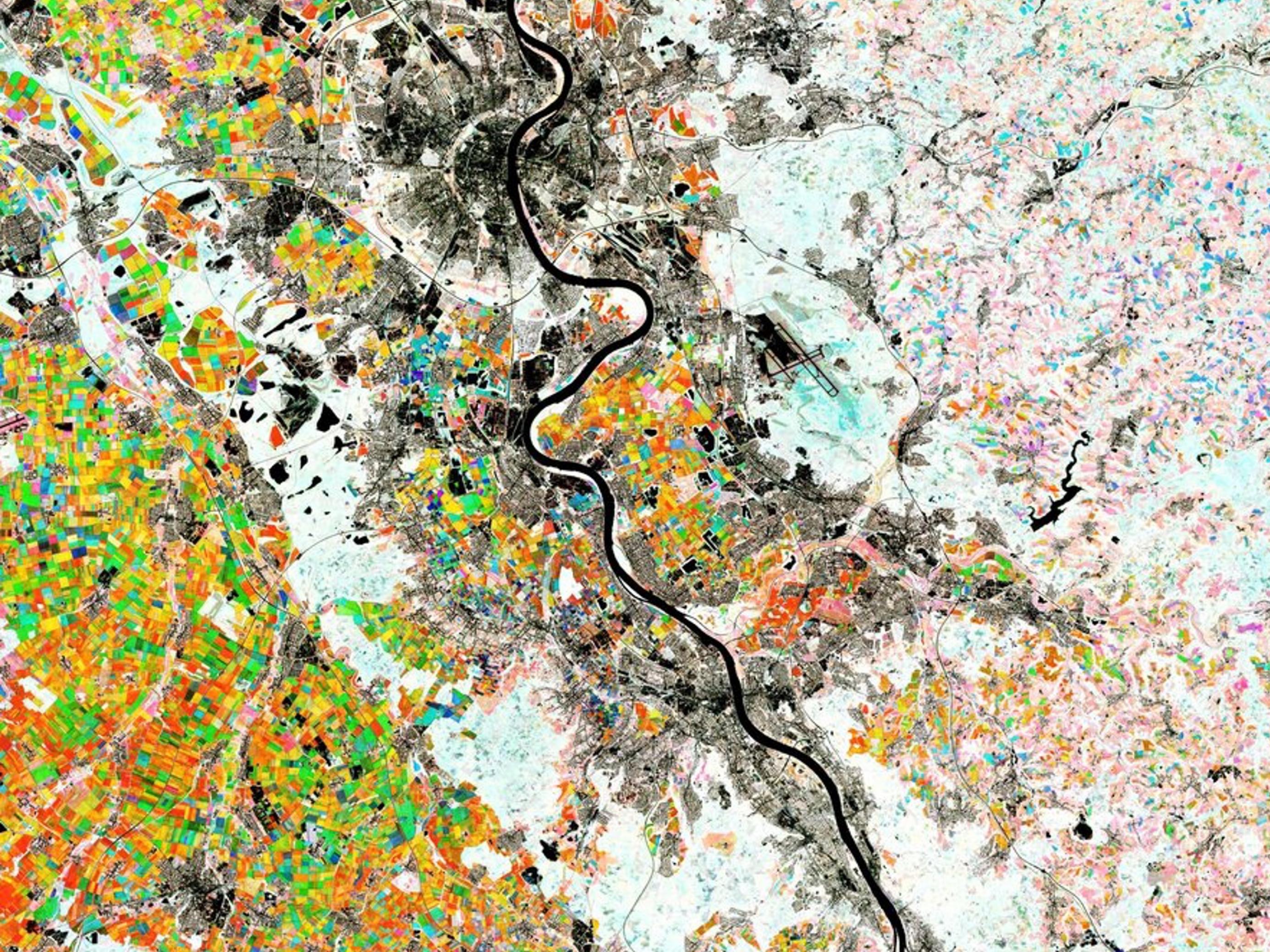 Luftbild mit dem Rhein als schwarzer, geschlängelter Linie, großen urbanen Flächen in grau und landwirtschaftlicher Nutzfläche in bunten Farben