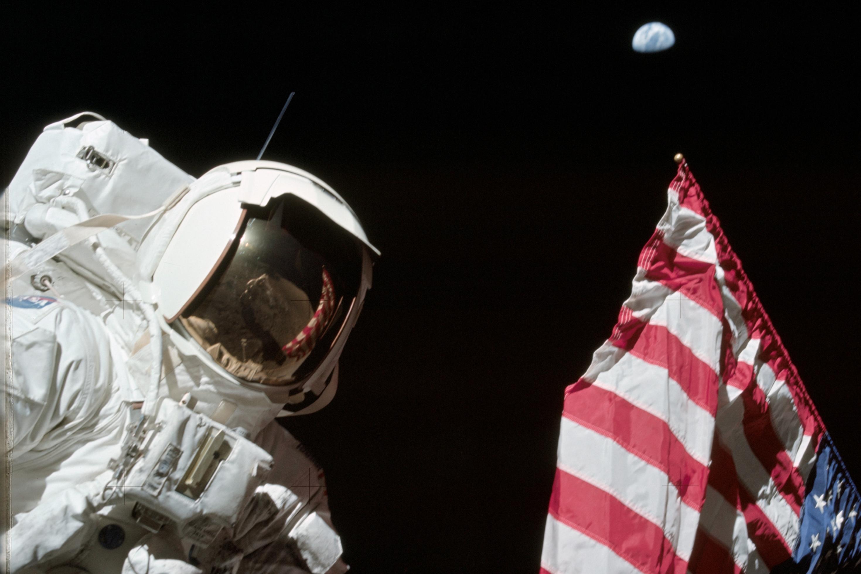 Schmitt steht am linken Bildrand, sein weißer Anzug und das rot-weiß-blau der Fahne bilden einen starken Kontrast zum Weltraum im Hintergrund.