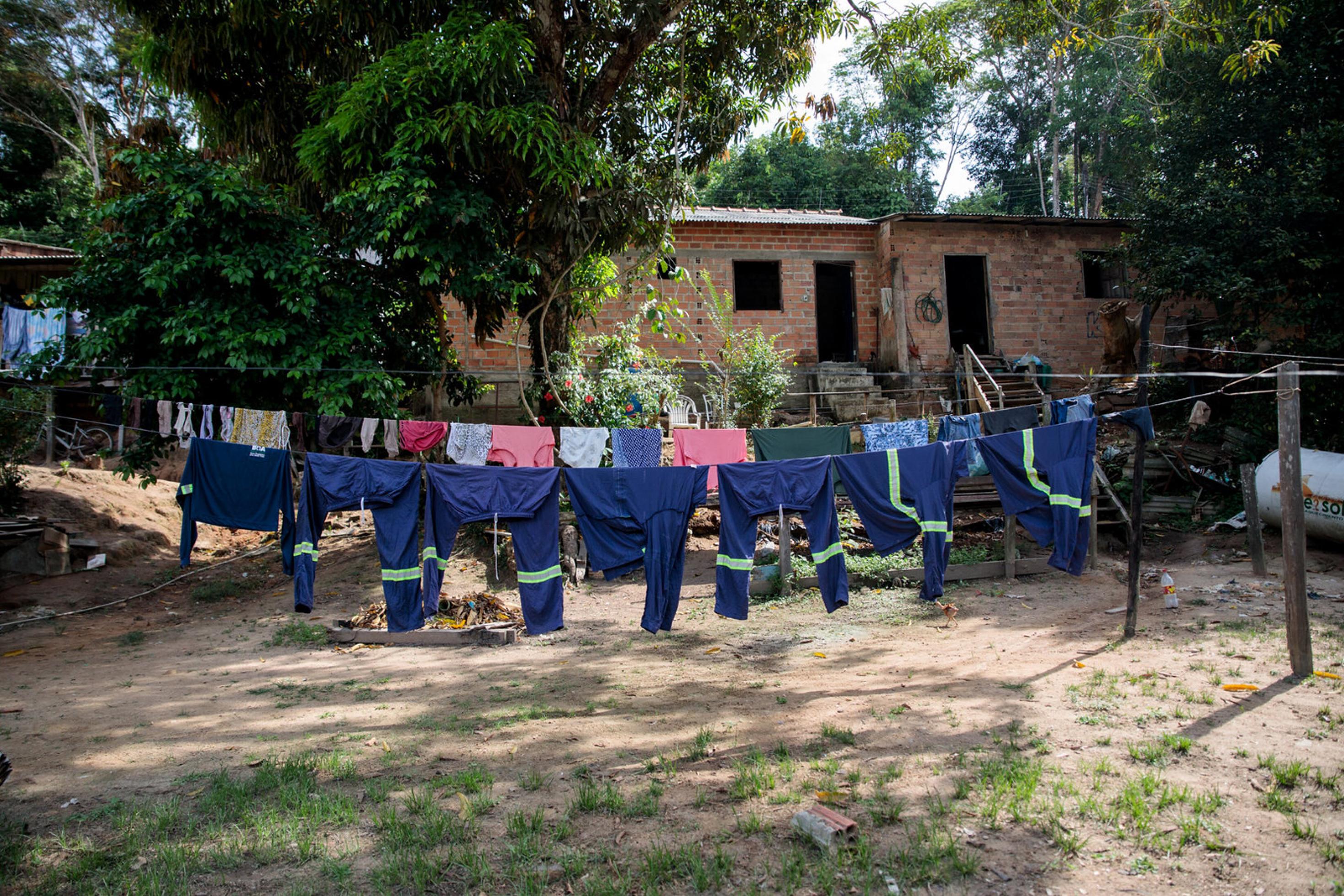 Ein Platz vor einem kleinen Häuschen: Auf einer Wäscheleine hängen viele dunkelblaue Uniformen von MRN-Arbeitern. Dahinter hängt Alltagskleidung.