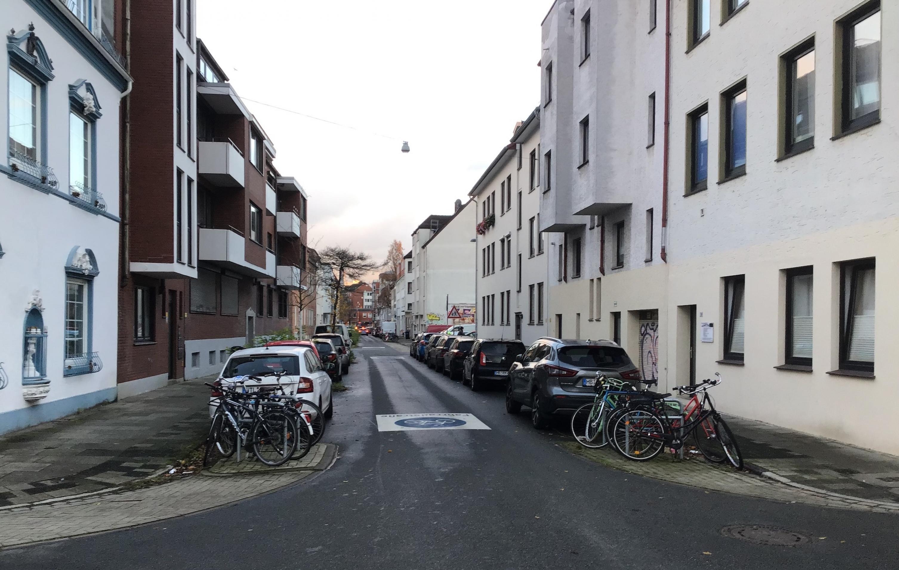 Direkt an der Kreuzung stehen Fahrräder, die an Bügeln angeschlossen sind. Hinter ihnen parken recht und links am Fahrbahnrand Autos