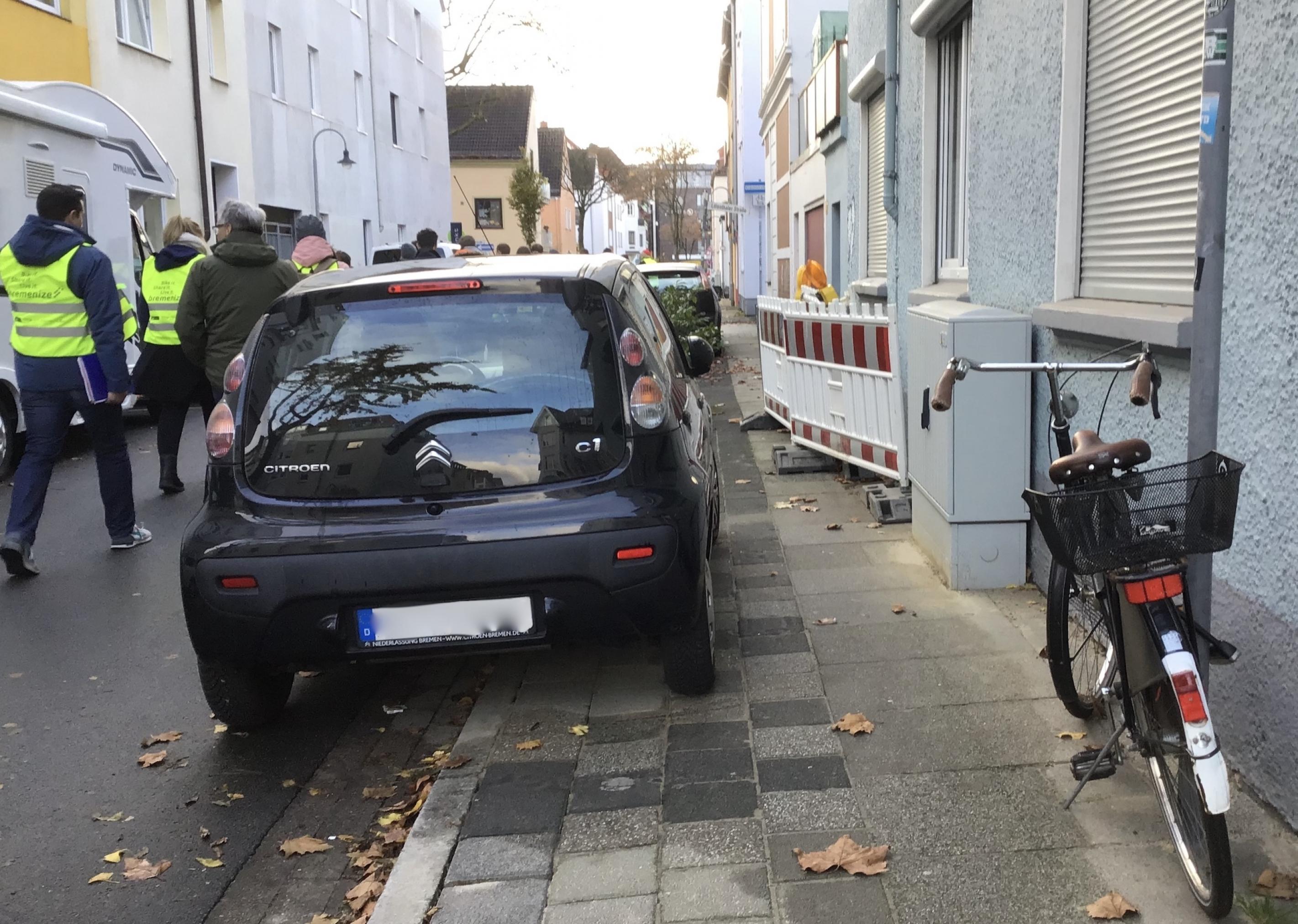 Der Gehweg wird versperrt von parkenden Autos und Baustellenbaken. Auf der Straße laufen Menschen