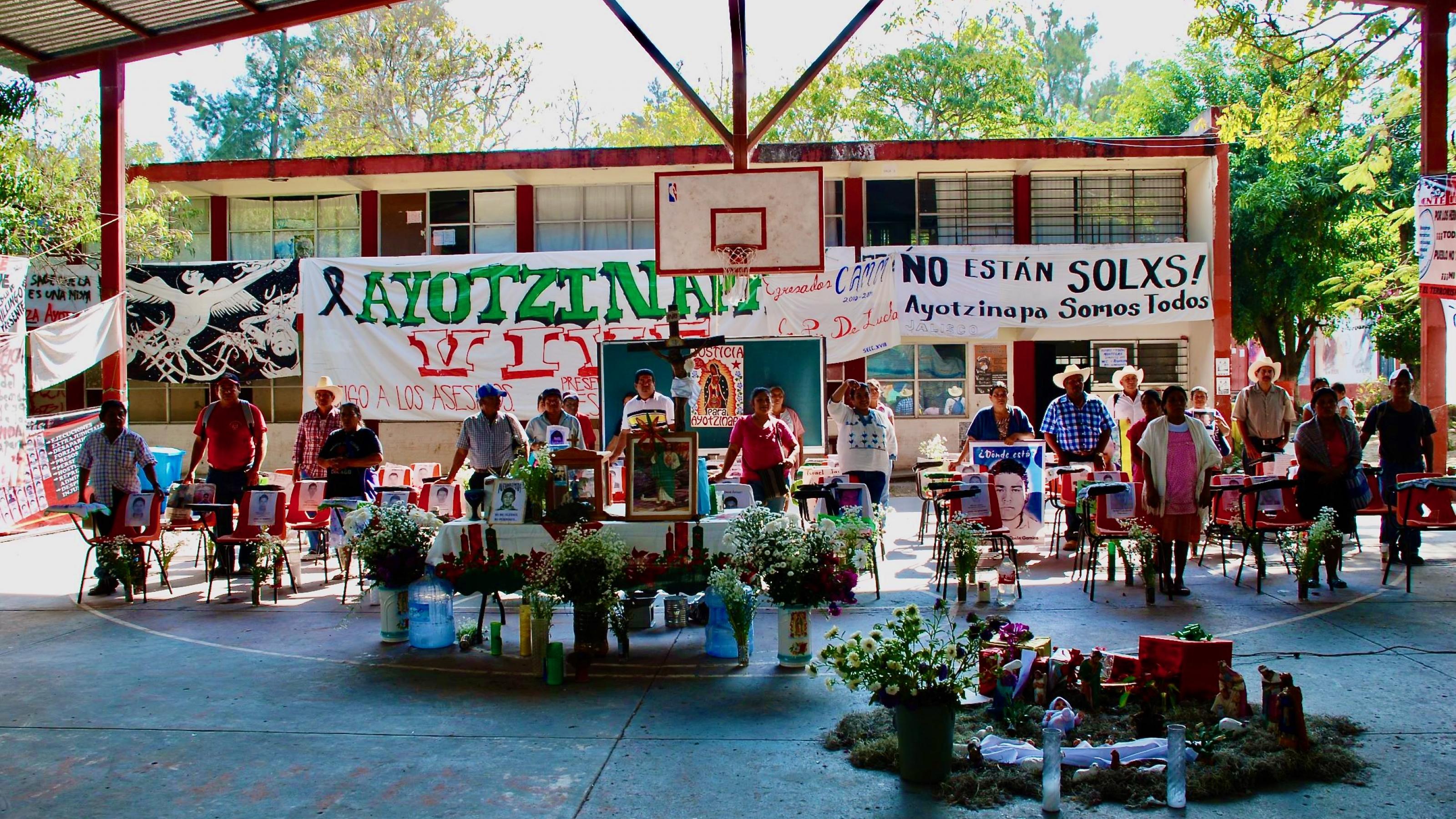 Menschen  stehen vor leeren Stühlen mit den Bildern ihrer verschwundenen Angehörigen im Lehrerseminar Ayotzinapa