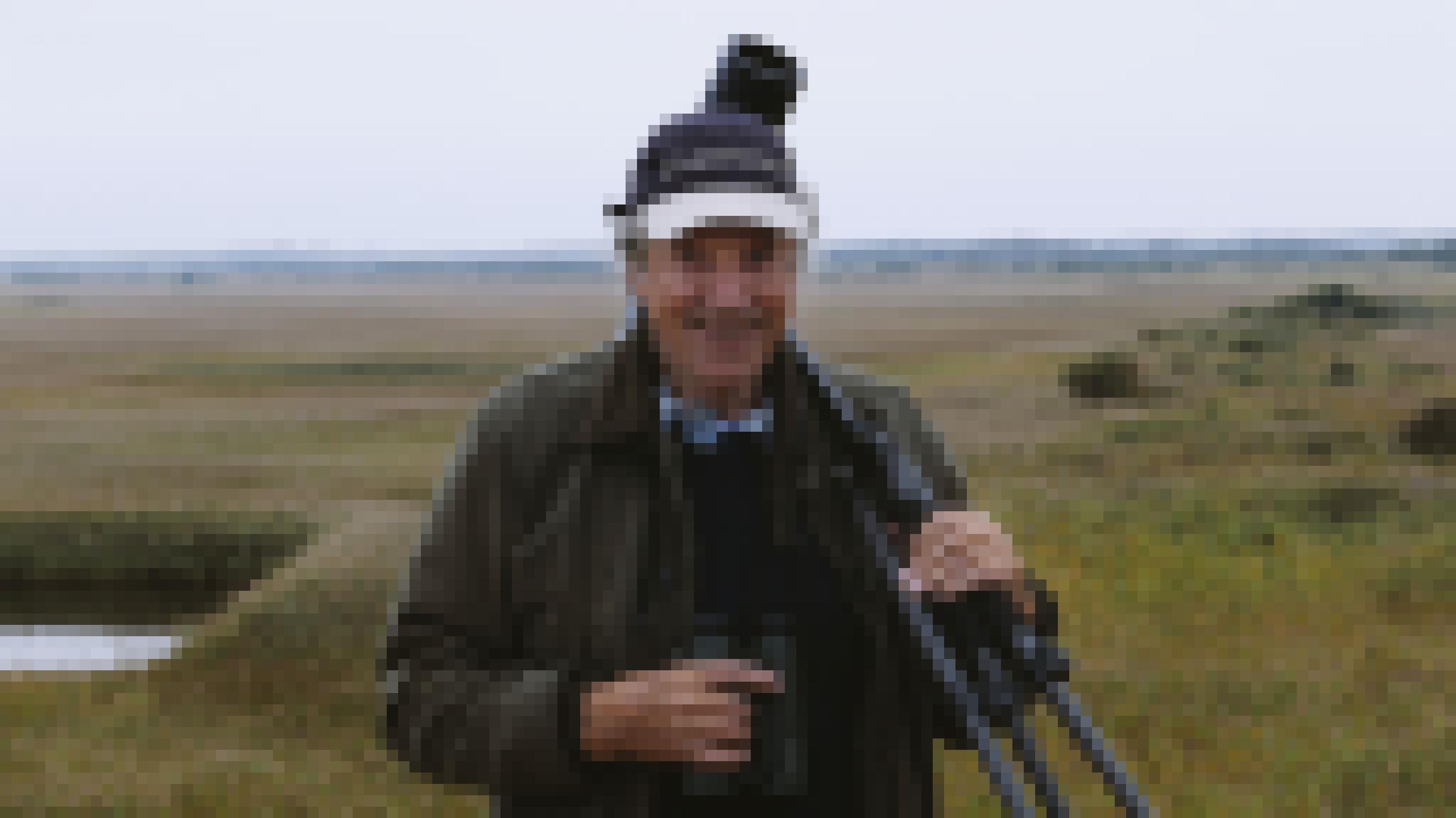 Ein älterer, freundlich lächelnder Herr steht mit Stativ über der Schulter in einer nebligen Landschaft.