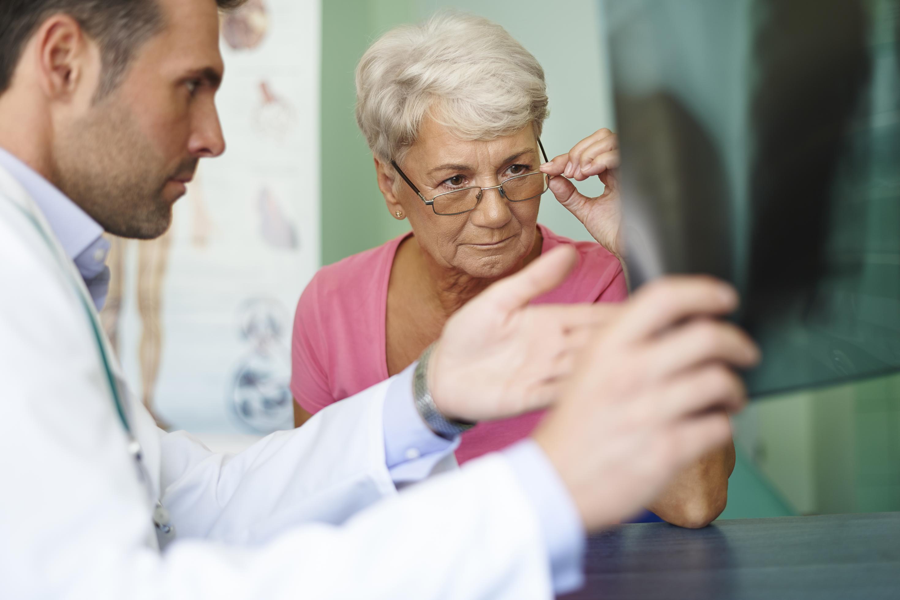 Ein Arzt zeigt einer Patientin ein Röntgenbild, die Patientin schiebt ihre Brille herunter, um besser sehen und verstehen zu können, was der Arzt erklärt
