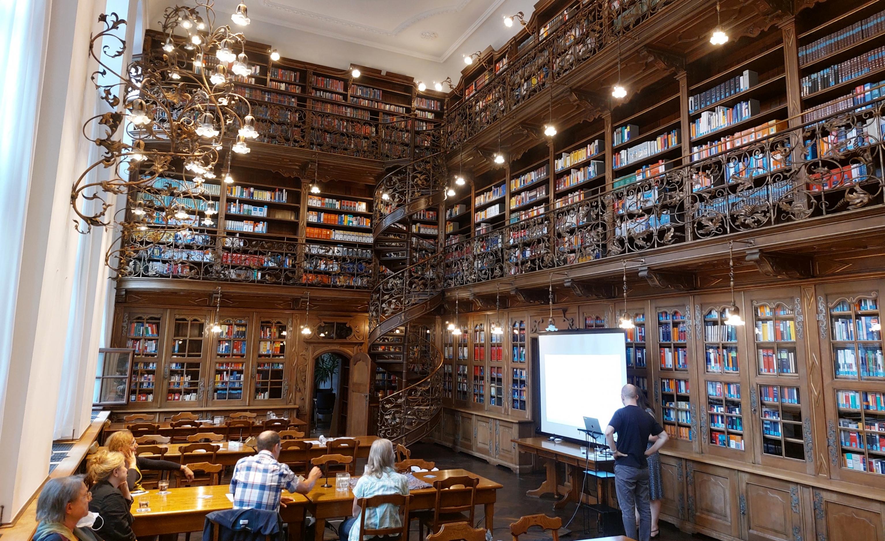 Historischer Lesesaal mit Holzregalen auf drei Ebenen, an Tischen mehrere Menschen, eine Leinwand.