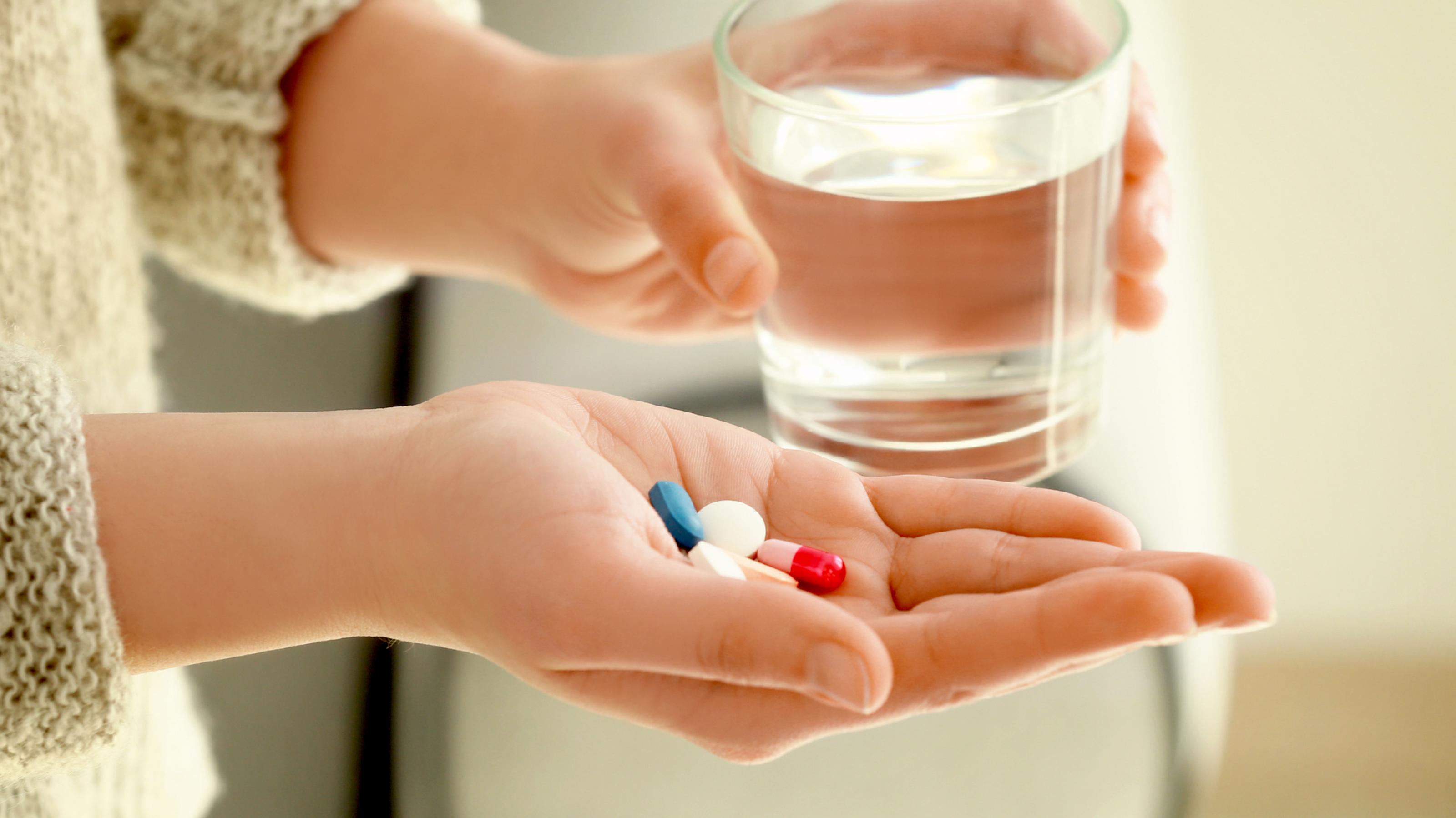 Eine geöffnete Hand hält mehrere Tabletten, eine zweite ein Glas Wasser.