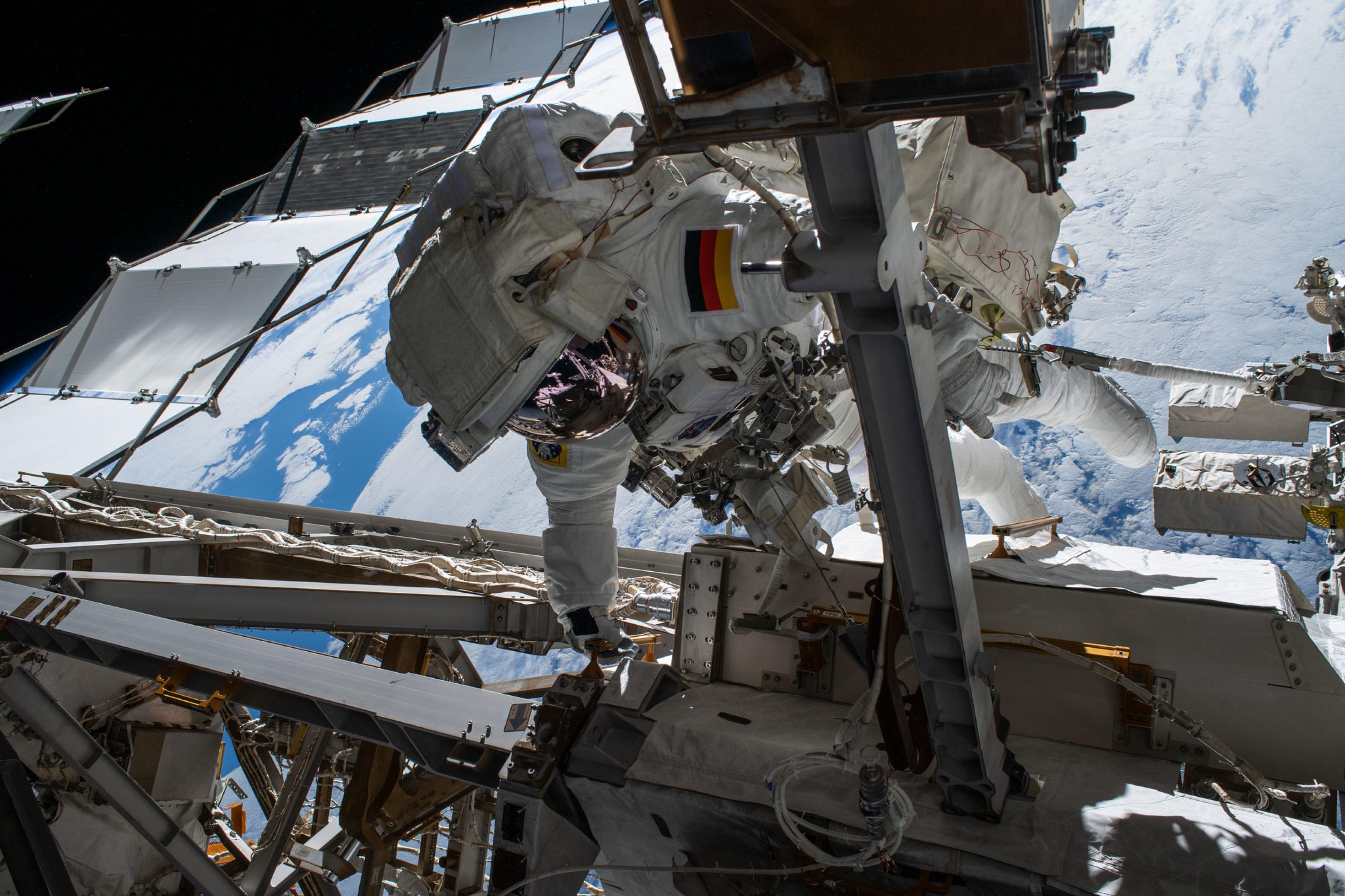 Ein Astronaut im Raumanzug hangelt sich an der Außenkonstruktion der Internationalen Raumstation entlang. Im Hintergrund ist ein Ausschnitt der wolkenbedeckten Erde zu sehen.