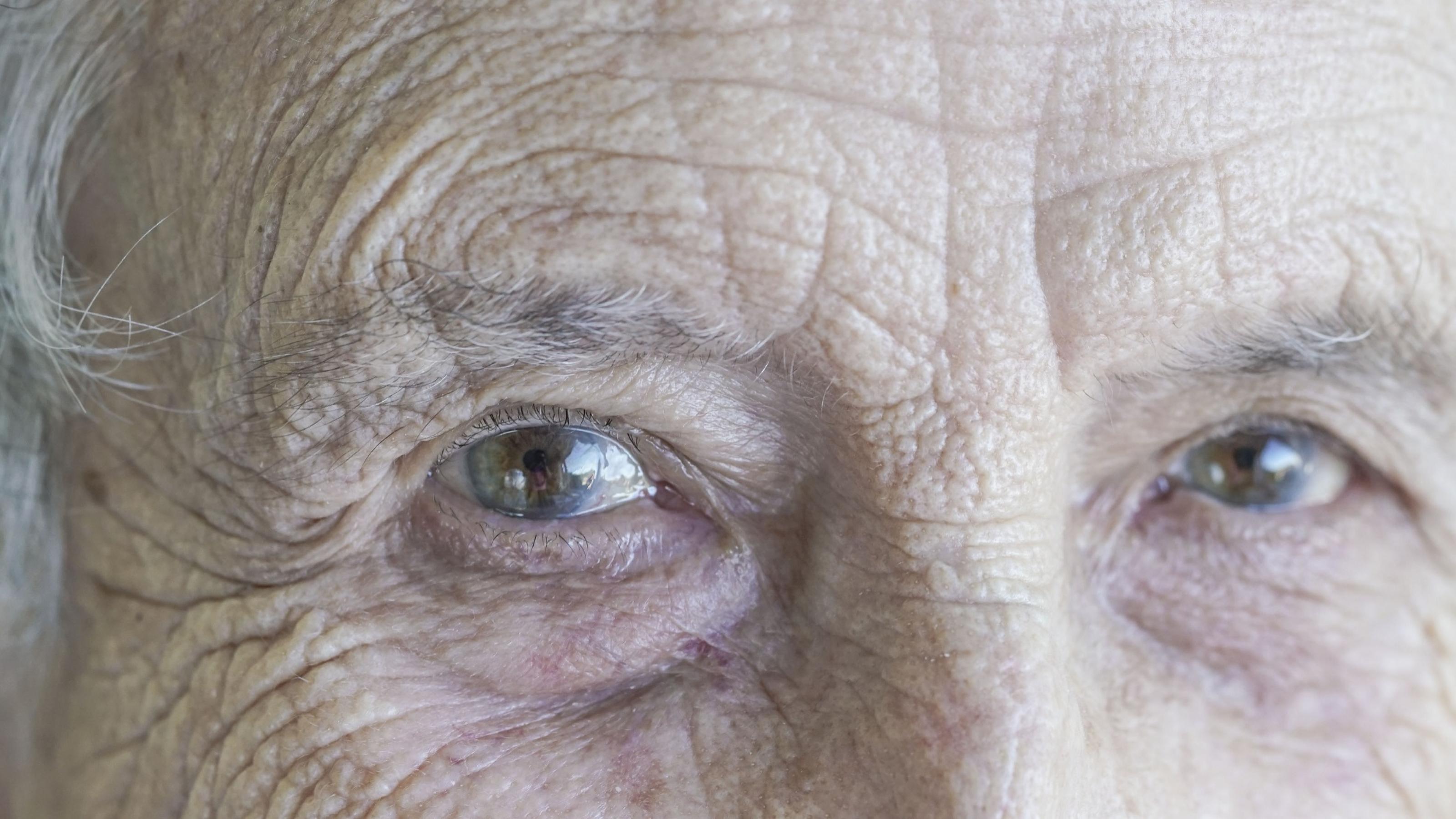 Eine Nahaufnahme zeigt die grau-braunen Augen einer alten Frau, die uns Betrachter direkt anschaut.