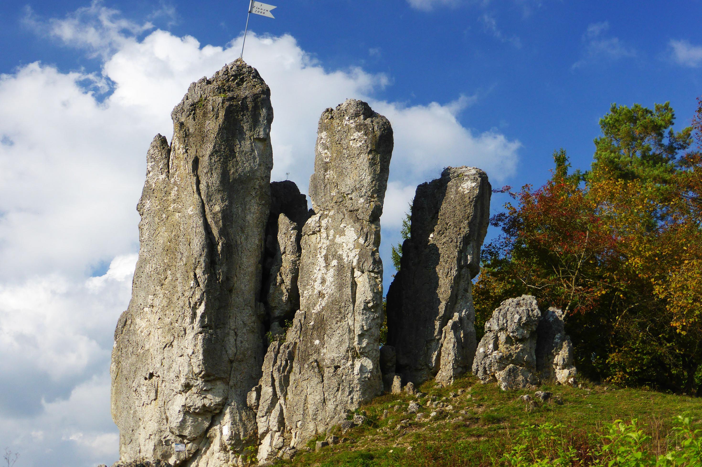 Diese Aufnahme der fränkischen drei Zinnen  zeigt auf der linken Zinnen einen Metallwimpel, im Hintergrund vor blauem Himmel dicke weiße Watte-Wolken.