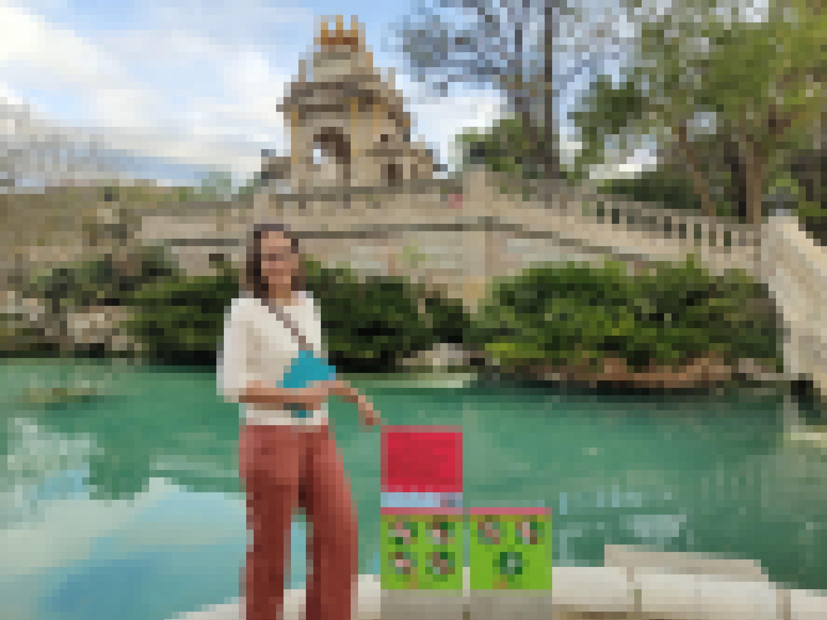 Eine Frau in roter Hose und weißem Pulli steht vor einem Springbrunnen in Barcelona, dessen Fontänen abgestellt sind. Das Becken ist mit Wasser gefüllt. Die Frau weist auf ein rotes Schild, auf dem in katalanisch steht, dass das Wasser nur dem Erhalt des Habitats der örtlichen Fauna dient.