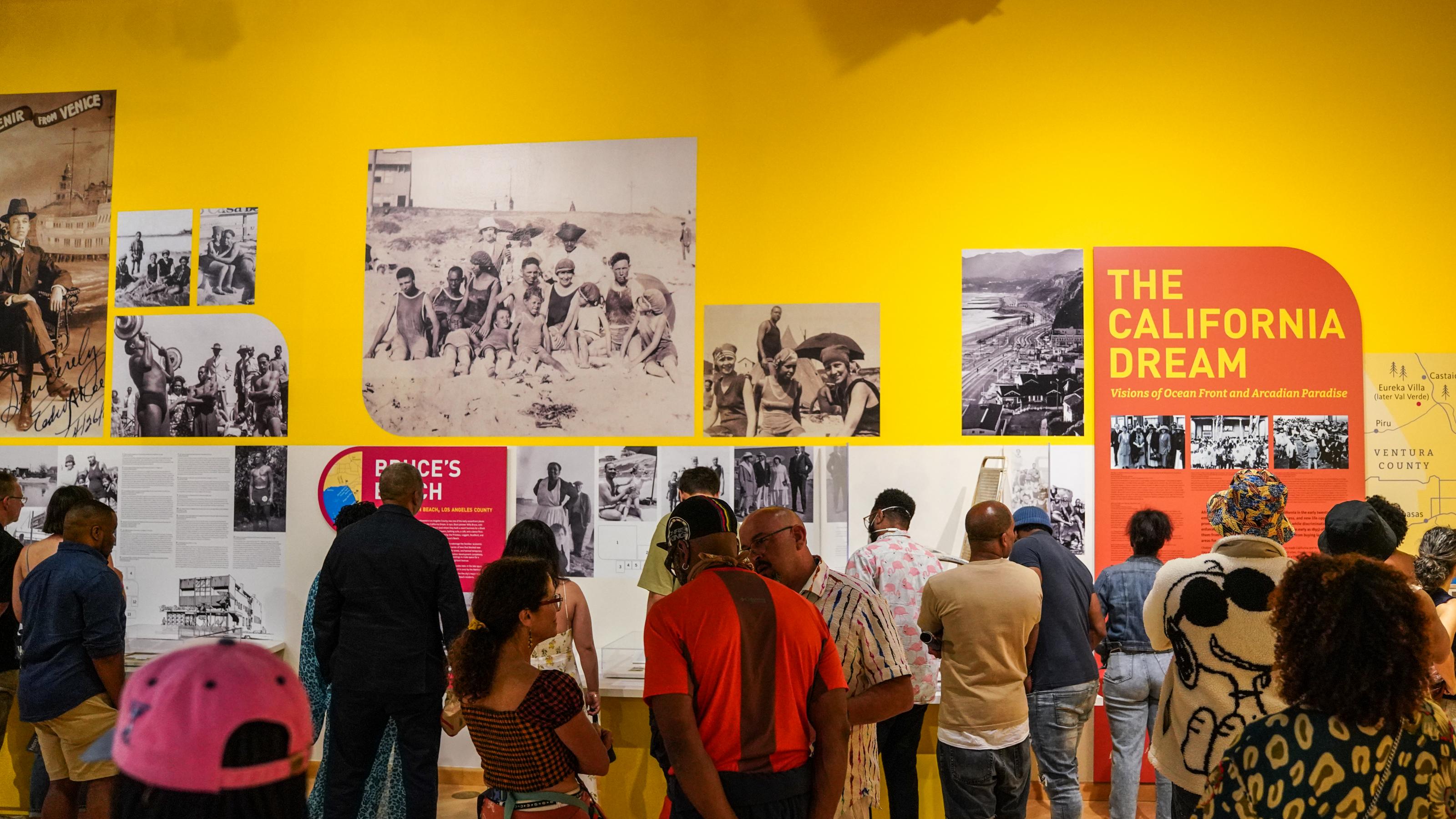 Ausstellungsbesucher betrachten eine gelbe Wand, die gefüllt ist mit schwarz-weiß Fotos aus dem Freizeitleben von Afroamerikanern. Rechts der Schriftzug: The California Dream.