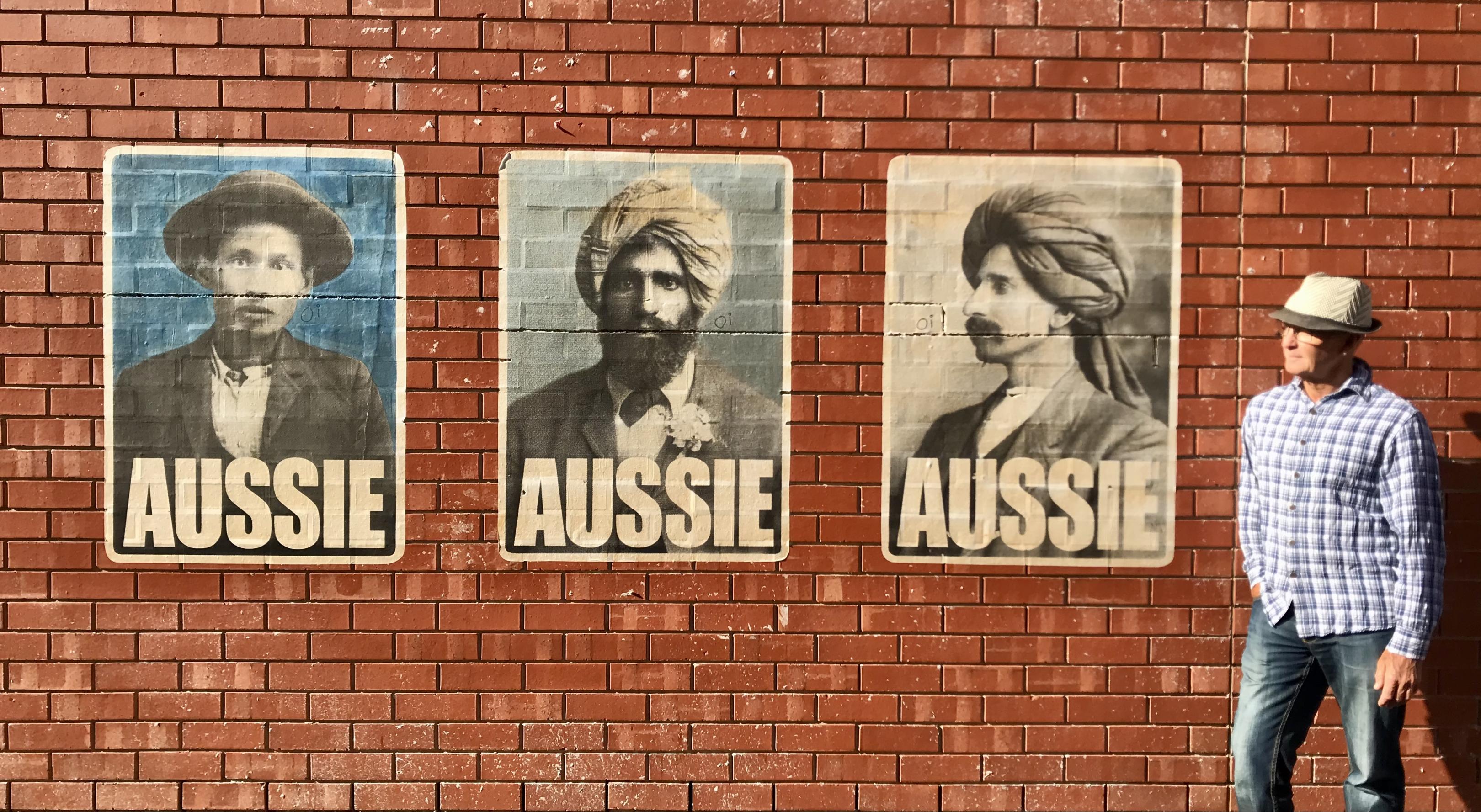 Ein Mann steht neben drei Postern an einer Hauswand, die jeweils den Titel  „Aussie“ tragen und Menschen mit unterschiedlicher kultureller Identität zeigen.