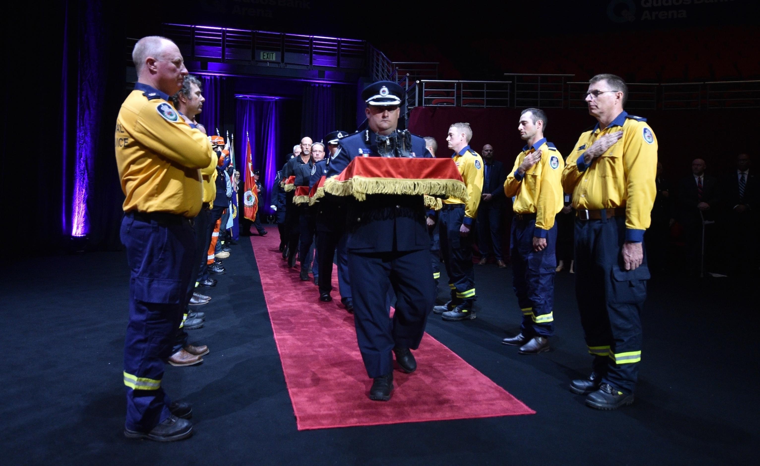 Feuerwehrmänner bilden in einer abgedunkelten Halle ein Spalier, während Kollegen in dekorierten Schachteln die Stiefel von Kameraden hereintragen, die bei den Bränden ums Leben gekommen sind, 