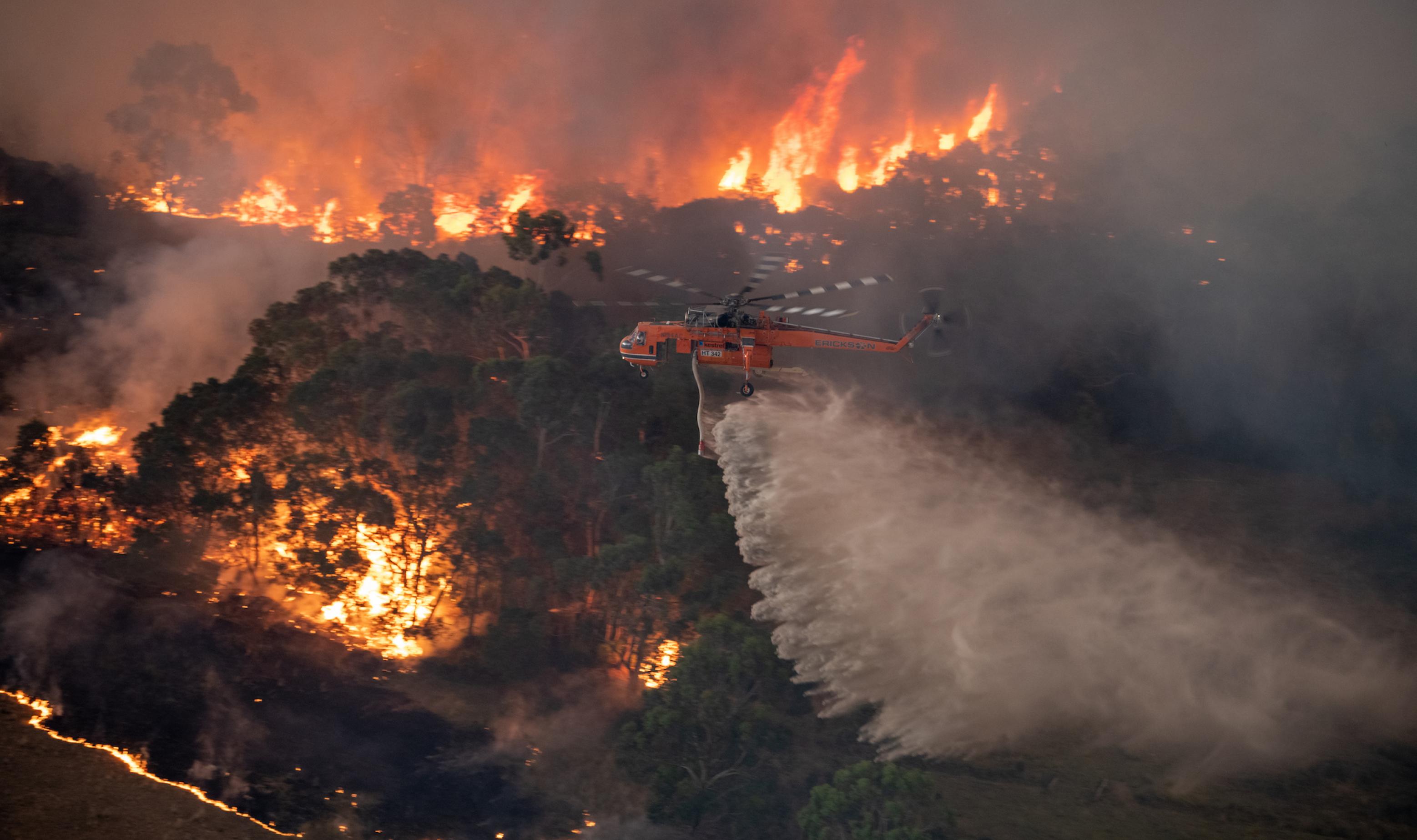 Das Bild zeigt, wie ein Feuerwehrhubschrauber bei Nacht eine große Menge Wasser über einem Waldbrand ausschüttet.