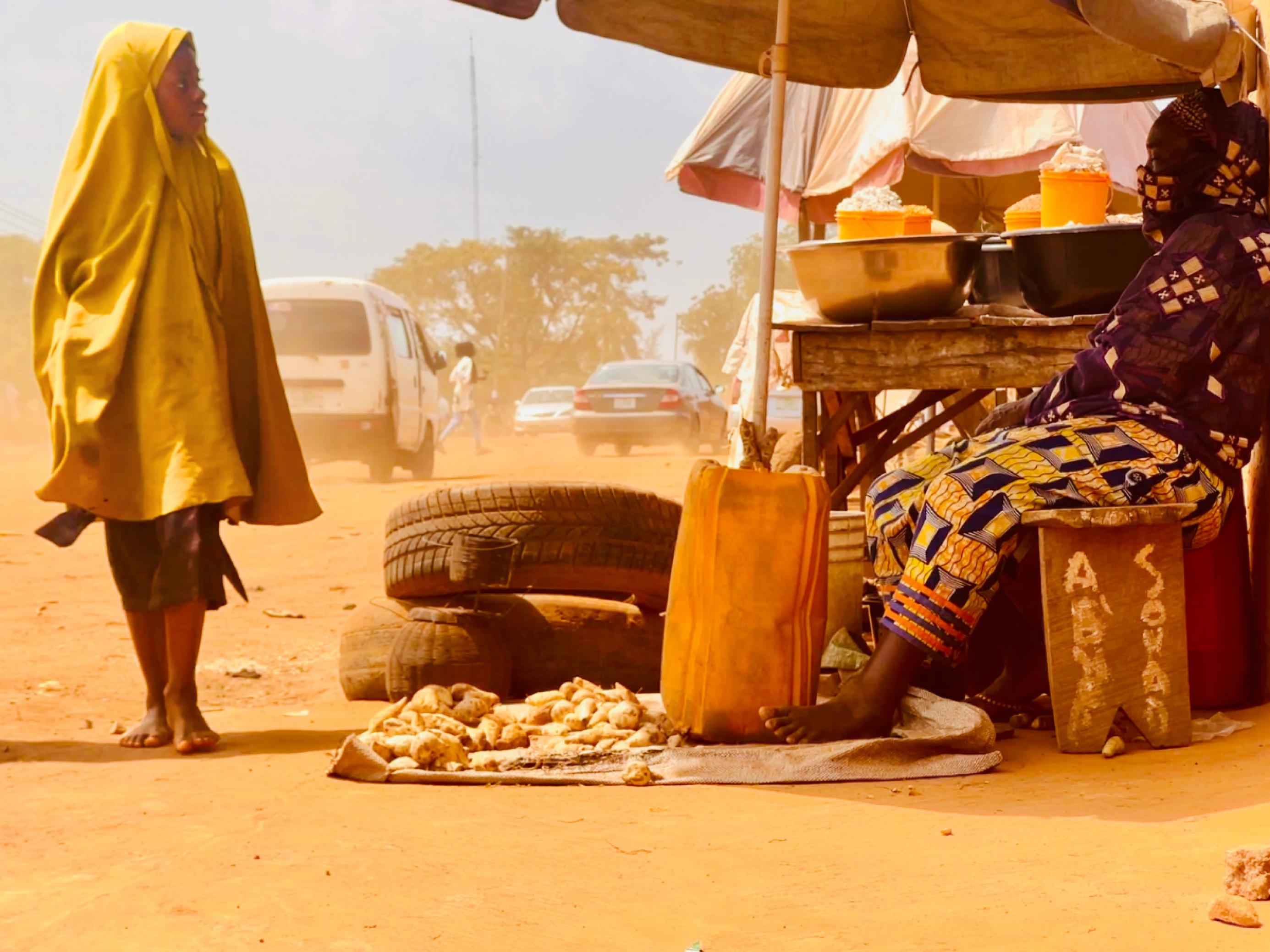 Ein Mädchen geht an einer Frau vorbei, die an einem staubigen Straßenrand in Kwara, Nigeria, vor kleinen Schüsseln sitzt und ihre Ernte zum Verkauf anbietet.