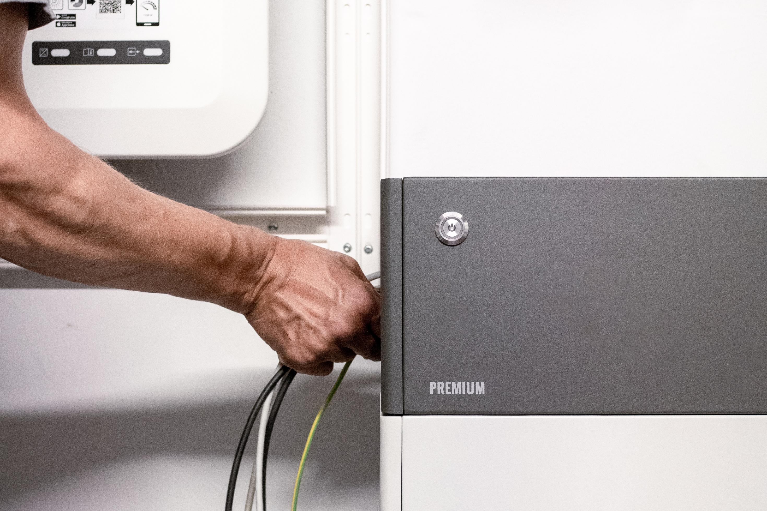 Eine Hand steckt ein Kabel in einen grau-weißen Batteriespeicher, im Hintergrund eine Elektro-Installation