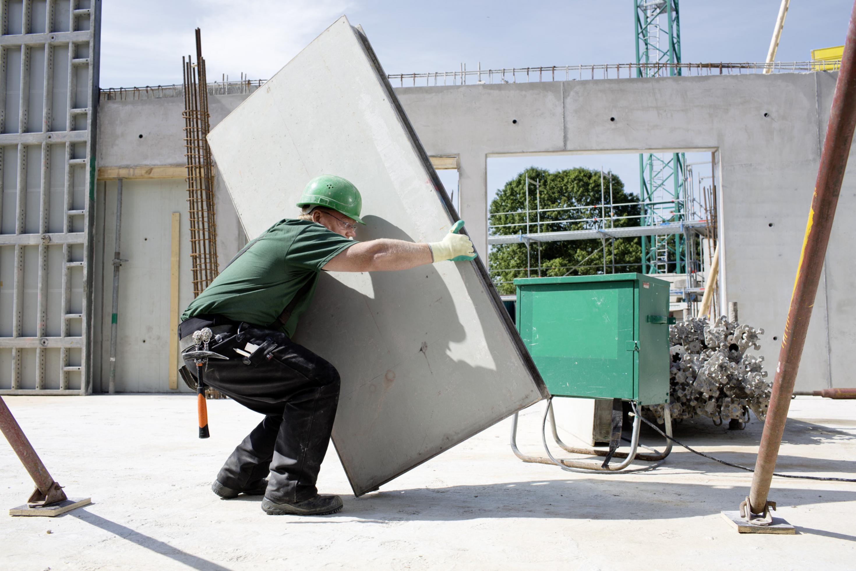 Ein Bauarbeiter mit grünem Helm ist in die Hocke gegangen und hält mit ausgestreckten Armen einen circa 1,5 mal 0,8 Meter großen Metallkasten. Die Umgebung ist der Rohbau eines größeren Gebäudes.