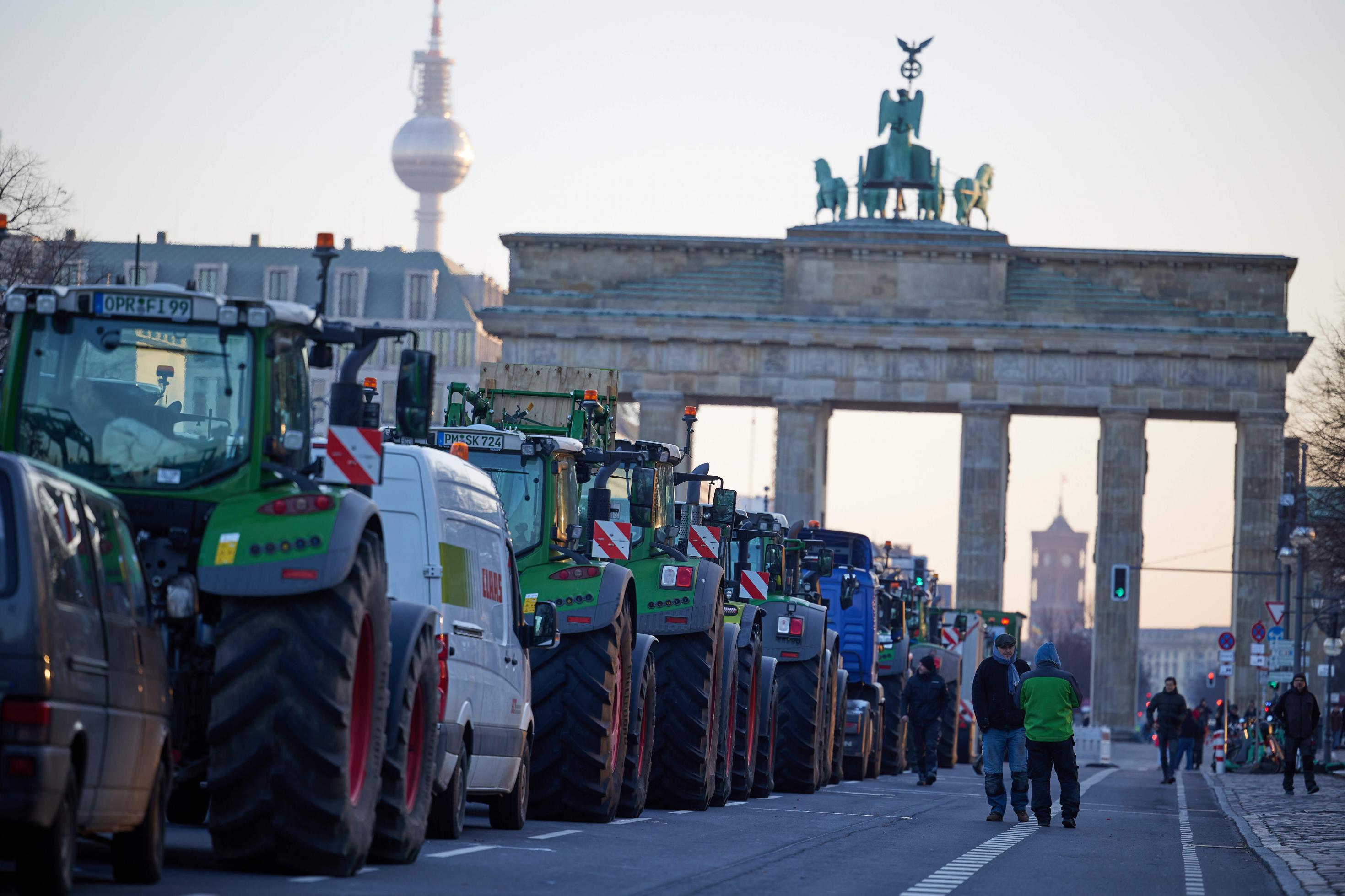 Halbtotale: Zahlreiche Traktoren - von hinten zu sehen - stehen bei einem Bauernprotest auf der Straße des 17. Juni vor dem Brandenburger Tor. Drei Bauern stehen in der Unschärfe neben den Traktoren. Das Foto wurde im Januar 2024 aufgenommen.