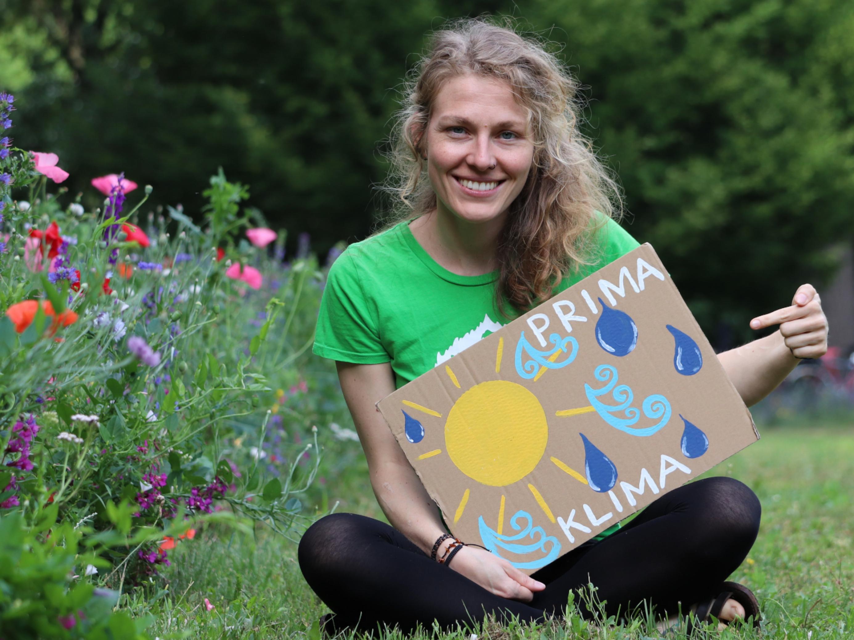 Frau Garske sitzt in einem blühenden Garten auf dem Rasen und hält ein Pappschild mit der Aufschrift „Prima Klima“